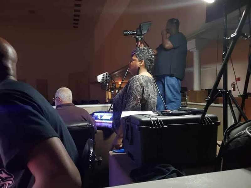 People seated in a room with a woman at a soundboard, a man with a camera, and equipment visible.
