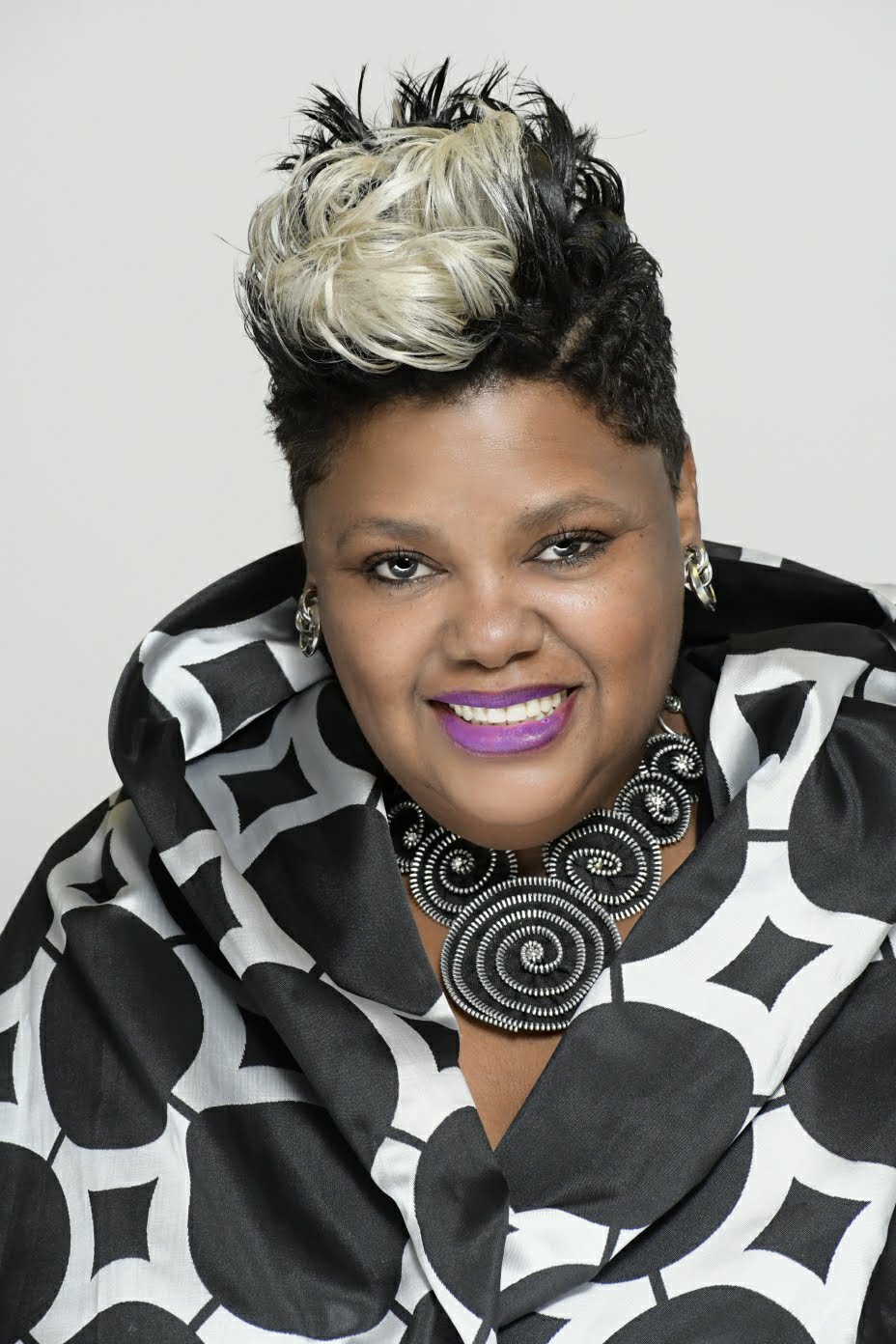 Woman with short, styled hair, wearing a black and white patterned outfit and a large spiral necklace, smiling.