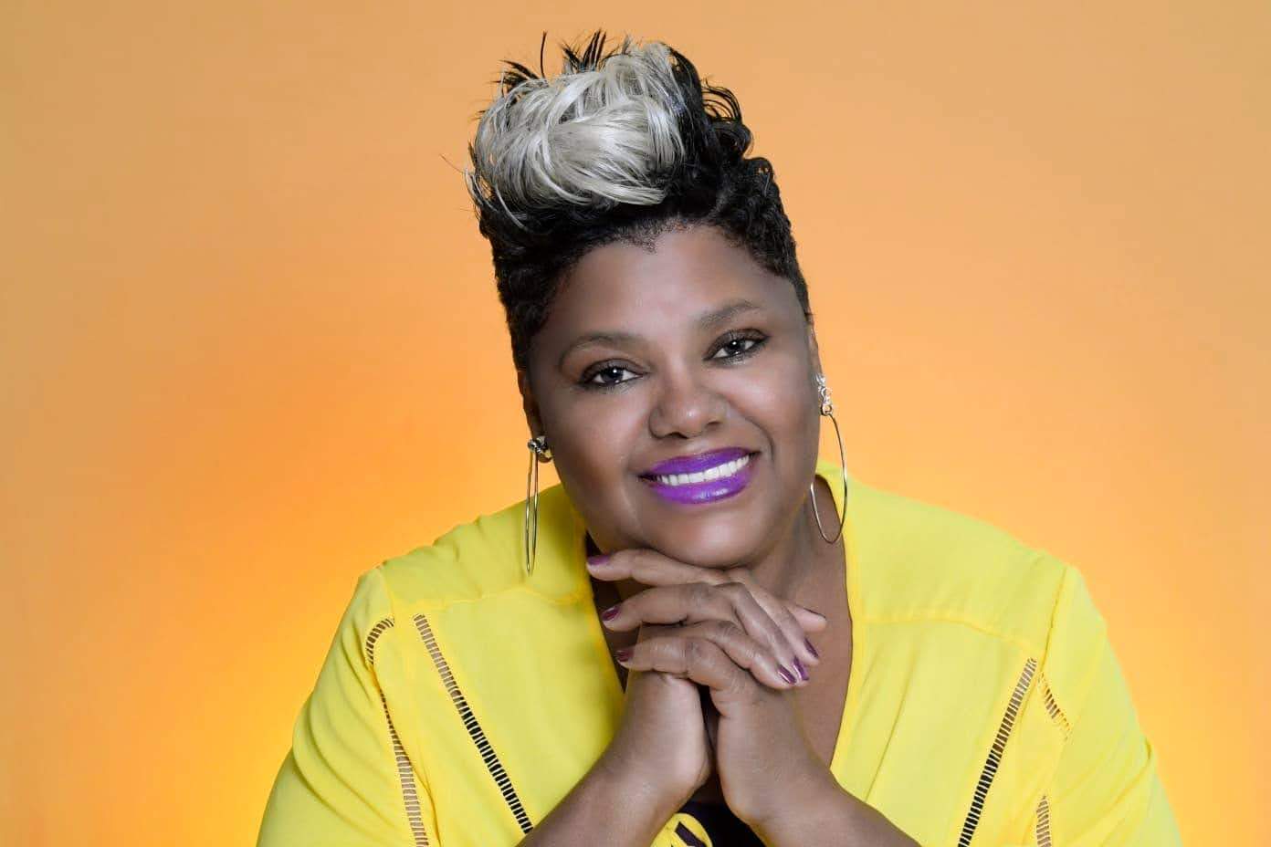 Woman with short, styled hair, wearing a yellow jacket, smiling with hands under chin, against a yellow-orange background.