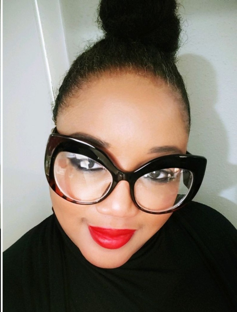 Close-up of a woman with glasses, red lipstick, and a high bun hairstyle, smiling at the camera.