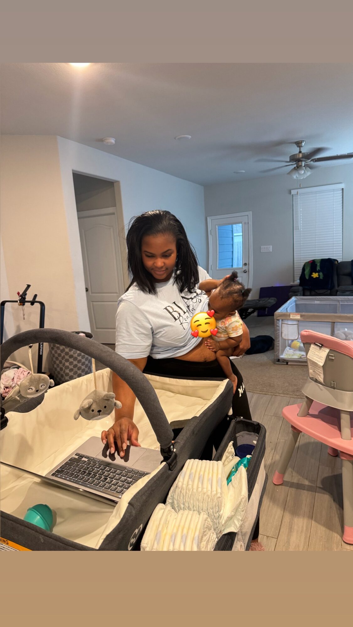 Woman holding a baby in a living room with a ceiling fan, door, and various furniture and items.