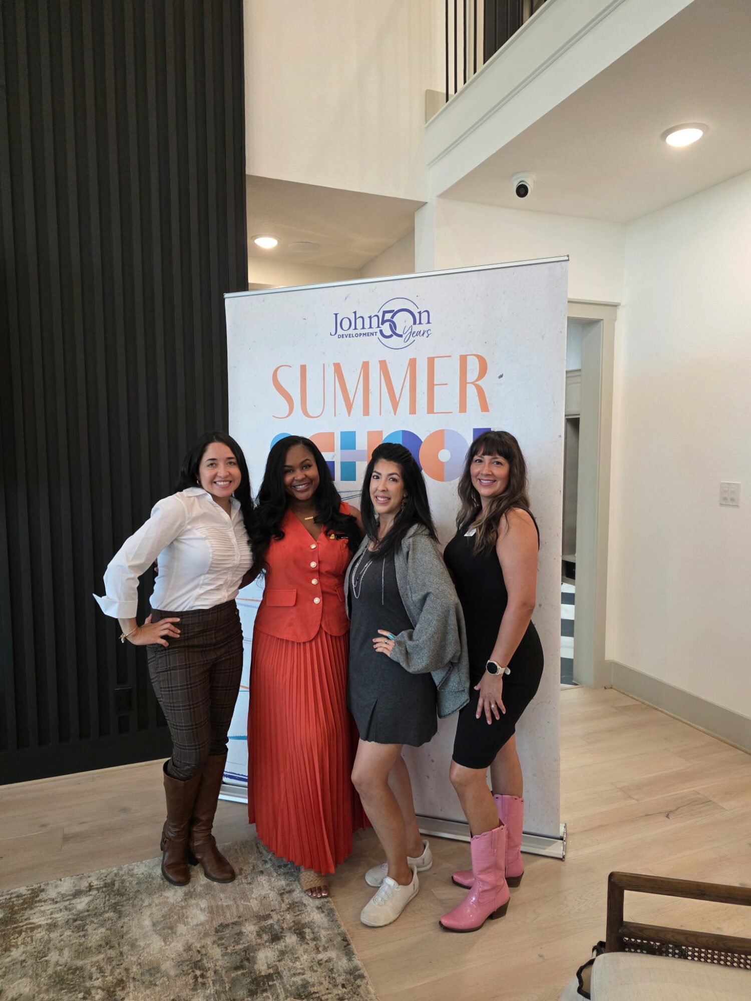 Four women standing together in front of a summer event sign indoors, smiling, with modern decor and a rug visible.
