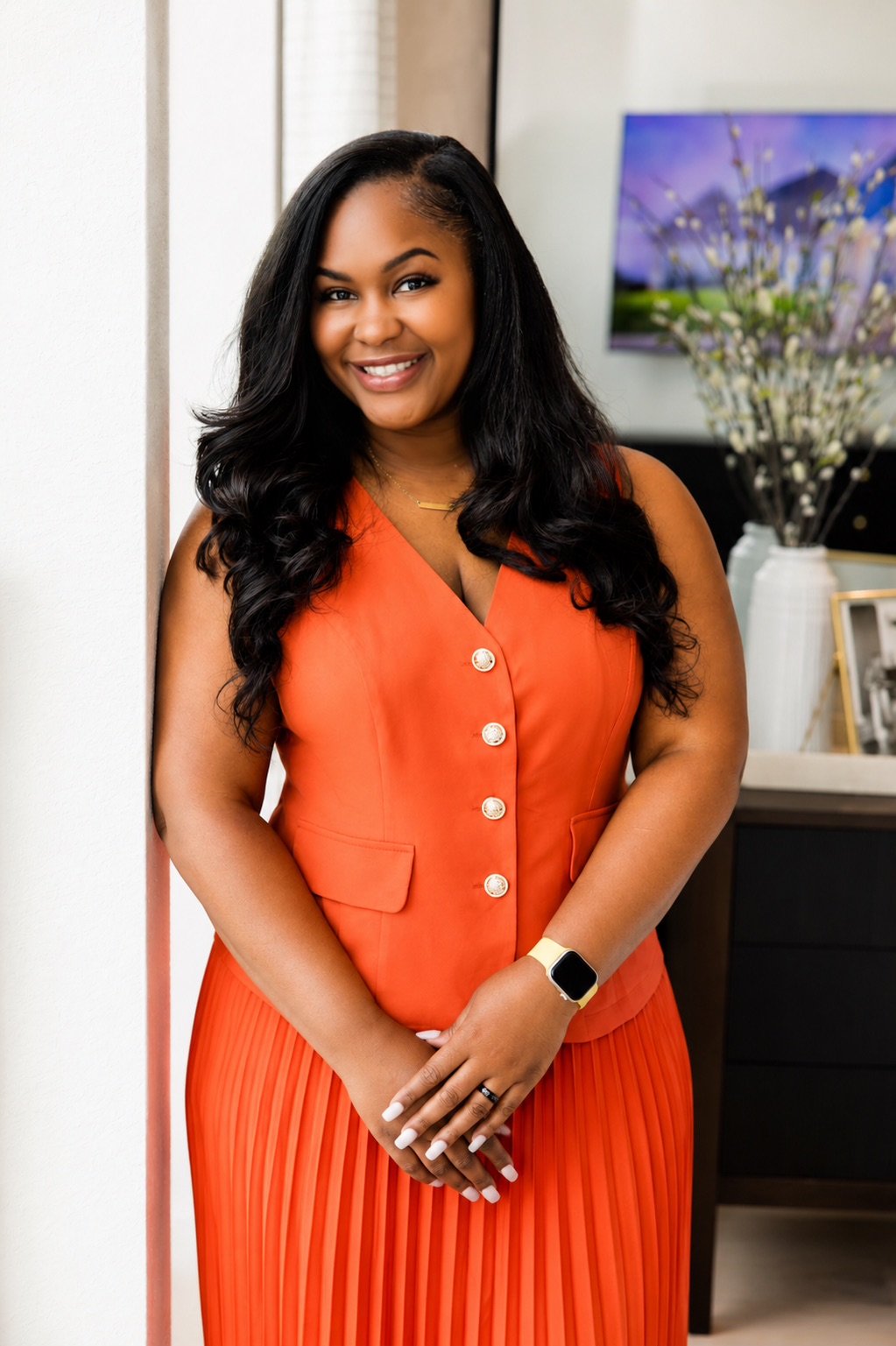 Smiling woman with long black hair in an orange sleeveless dress with buttons, standing indoors near a window and a TV.