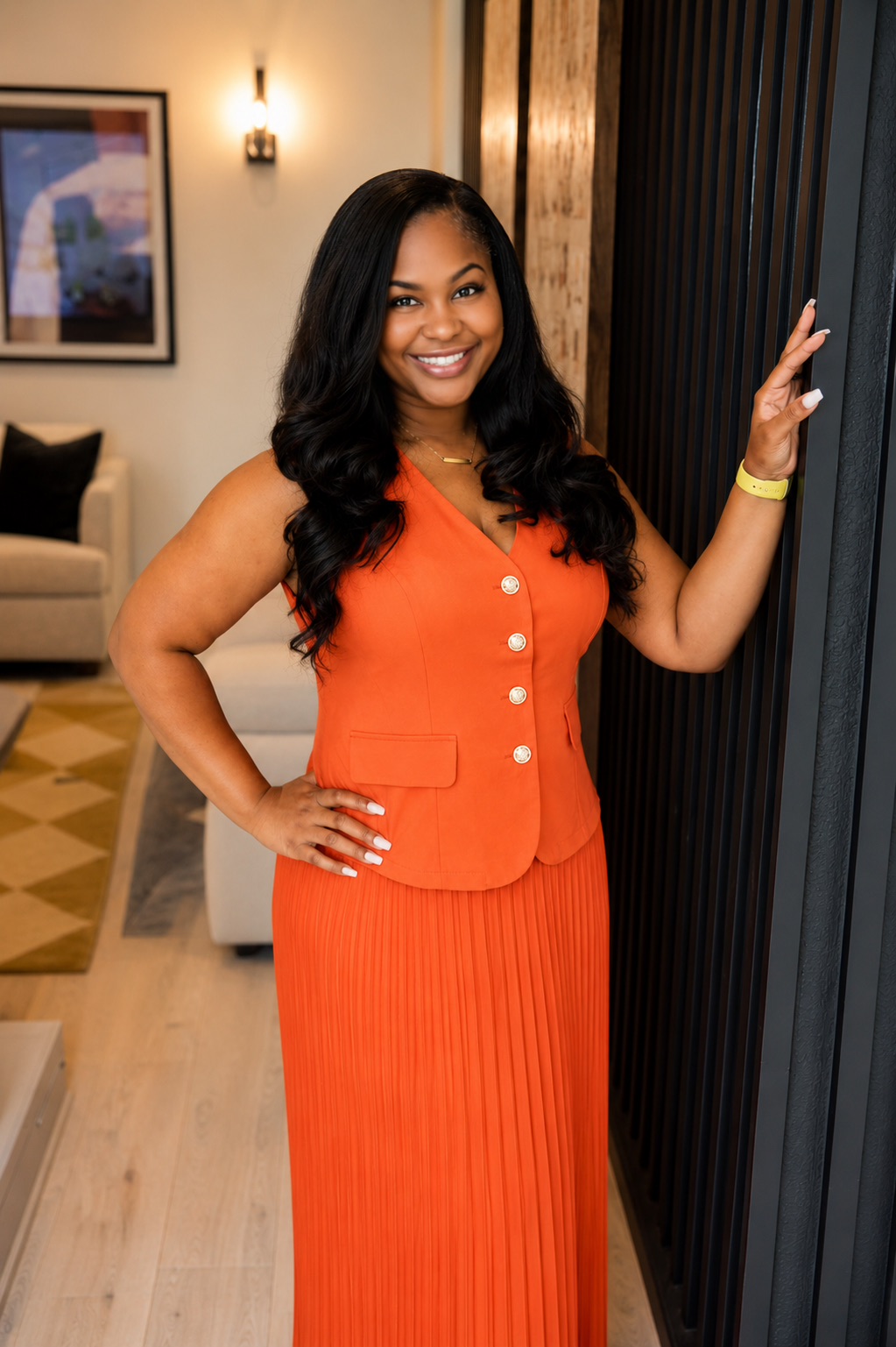 Woman in orange dress smiling, standing indoors near a dark wall, with a hand on the wall and a room in the background.