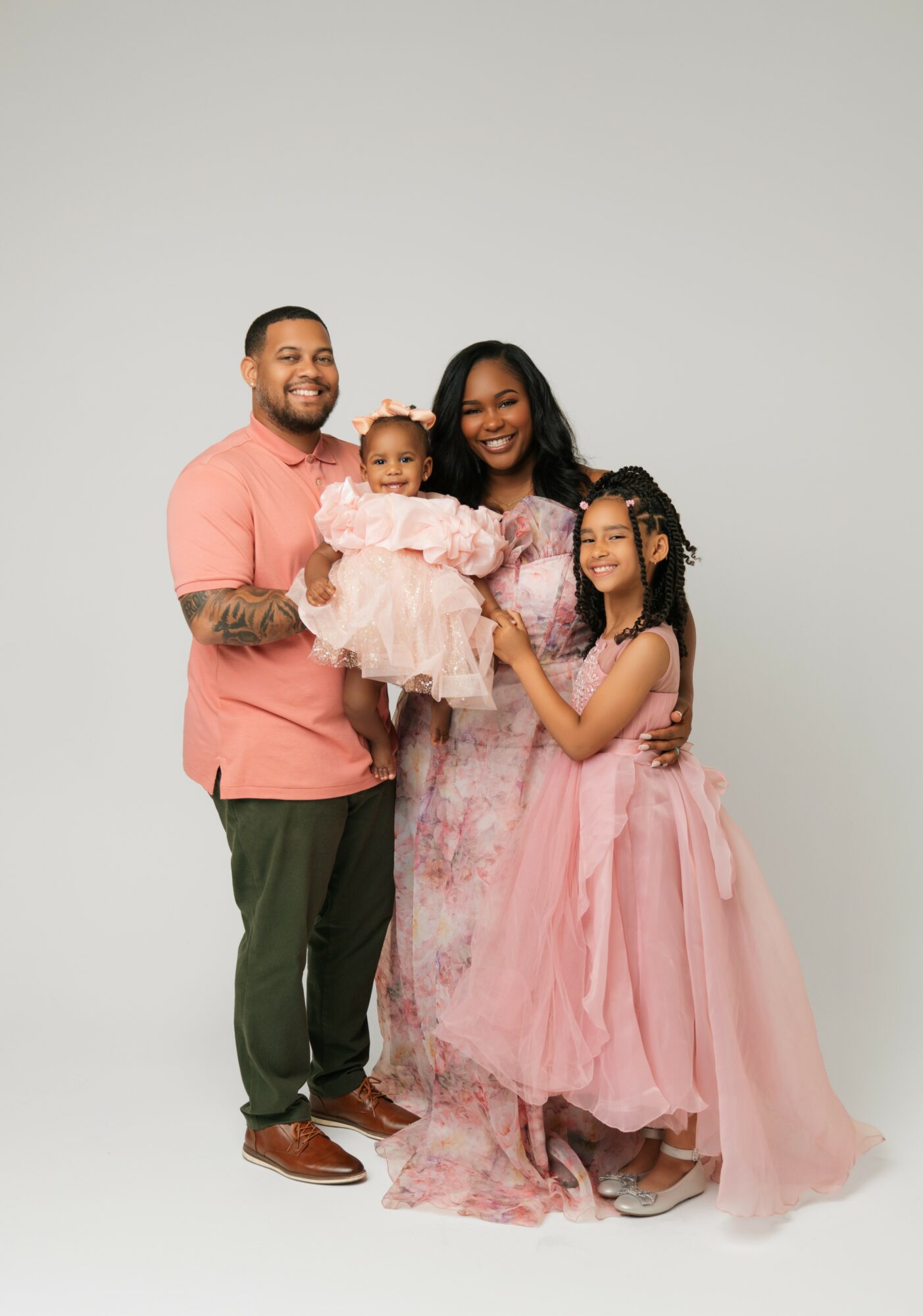 Family of four smiling, with a man, woman, and two girls, all dressed in pink, standing together against a plain background.