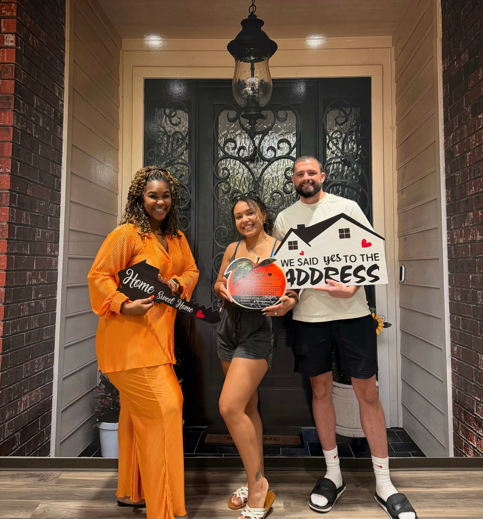 Three people standing on porch in front of door, holding signs, smiling, with hanging light above, brick walls on sides.