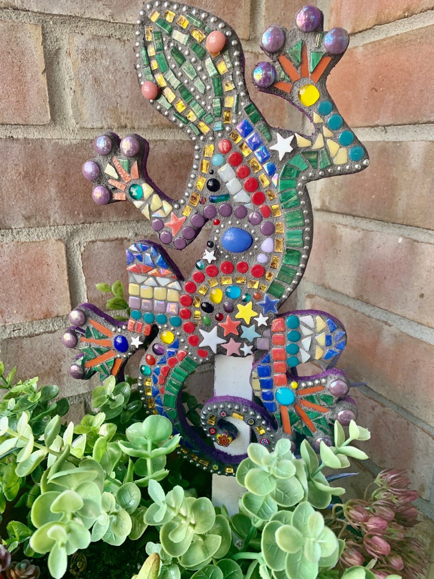 Colorful mosaic lizard sculpture with vibrant patterns, placed outdoors against a brick wall, surrounded by green plants.