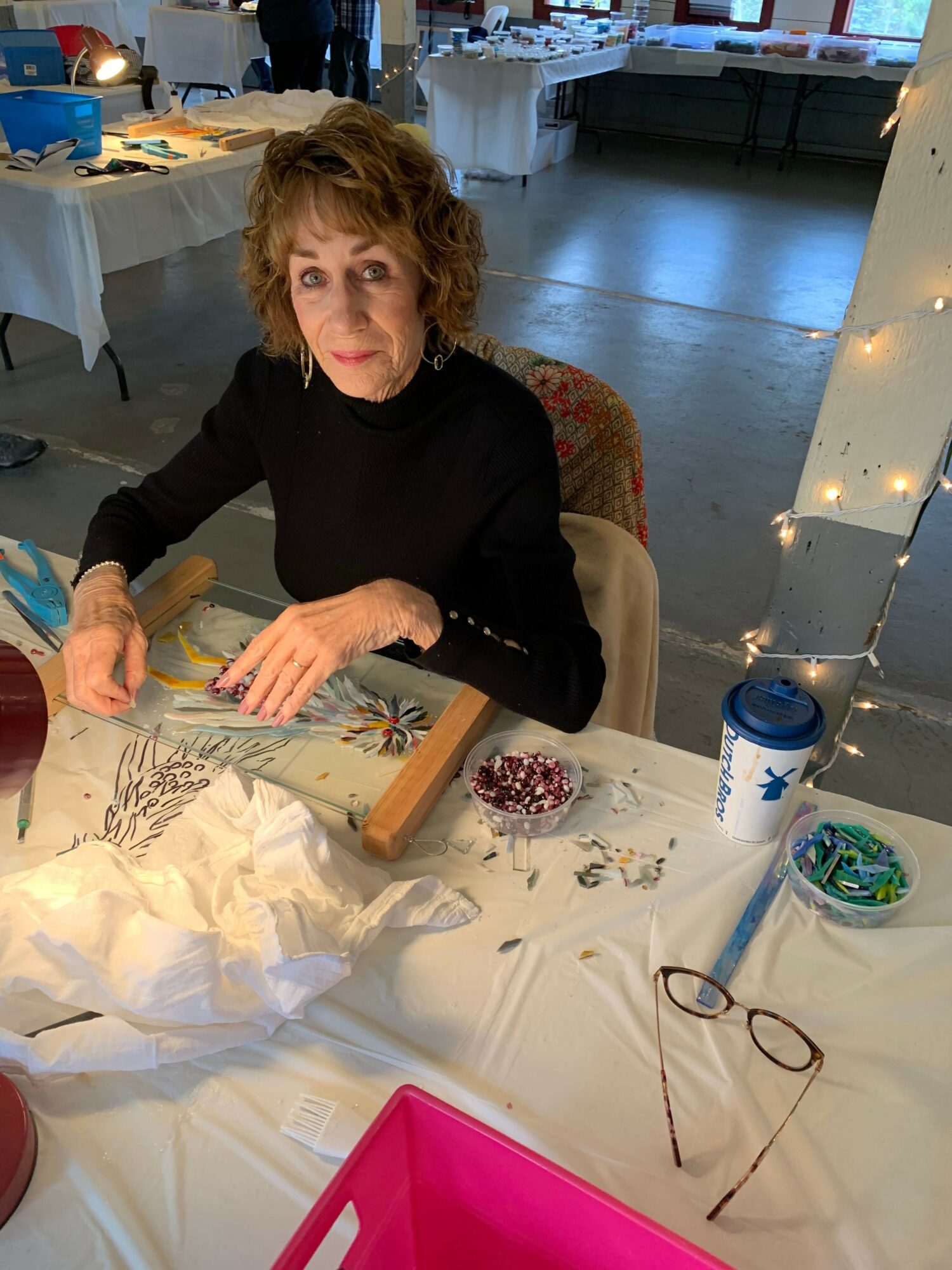 Woman with curly hair in black shirt working on craft project at table with supplies, pink container, and string lights.
