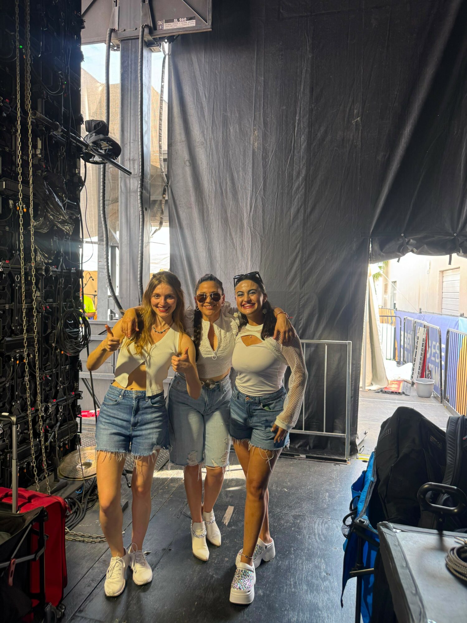 Three women standing together, smiling, giving thumbs up, in a backstage area with equipment and curtains.