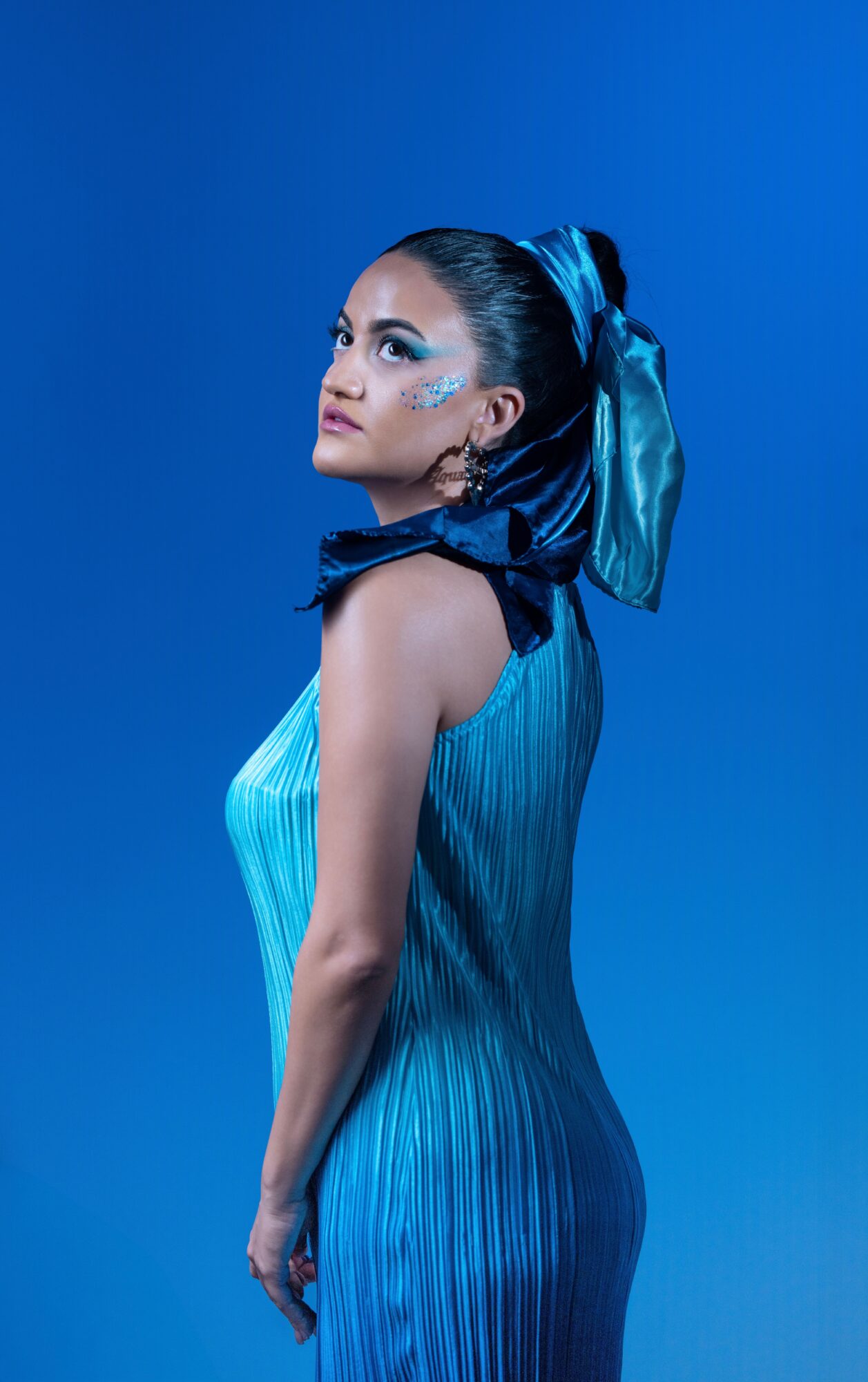 Woman with makeup and a large blue bow in her hair, wearing a blue dress, against a blue background.