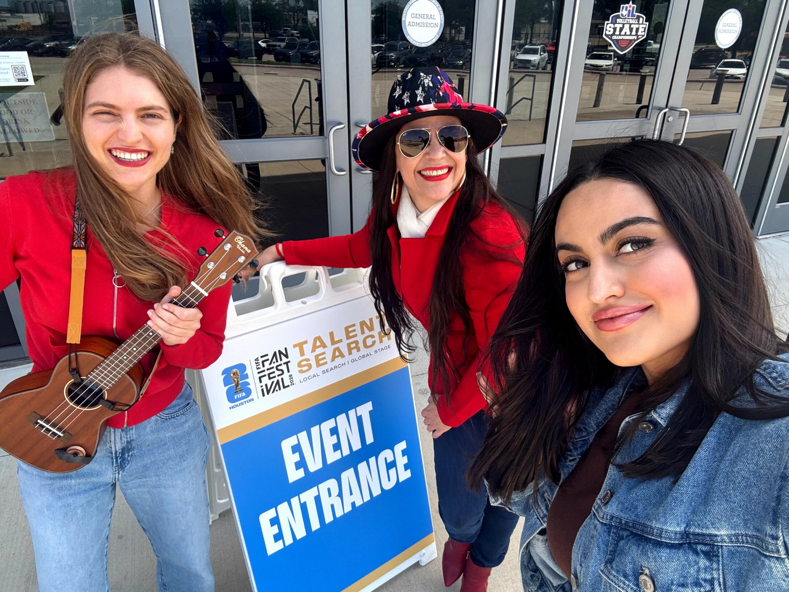 Three women smiling, one holding a ukulele, standing outside a building with an event entrance sign, all wearing colorful clothing.
