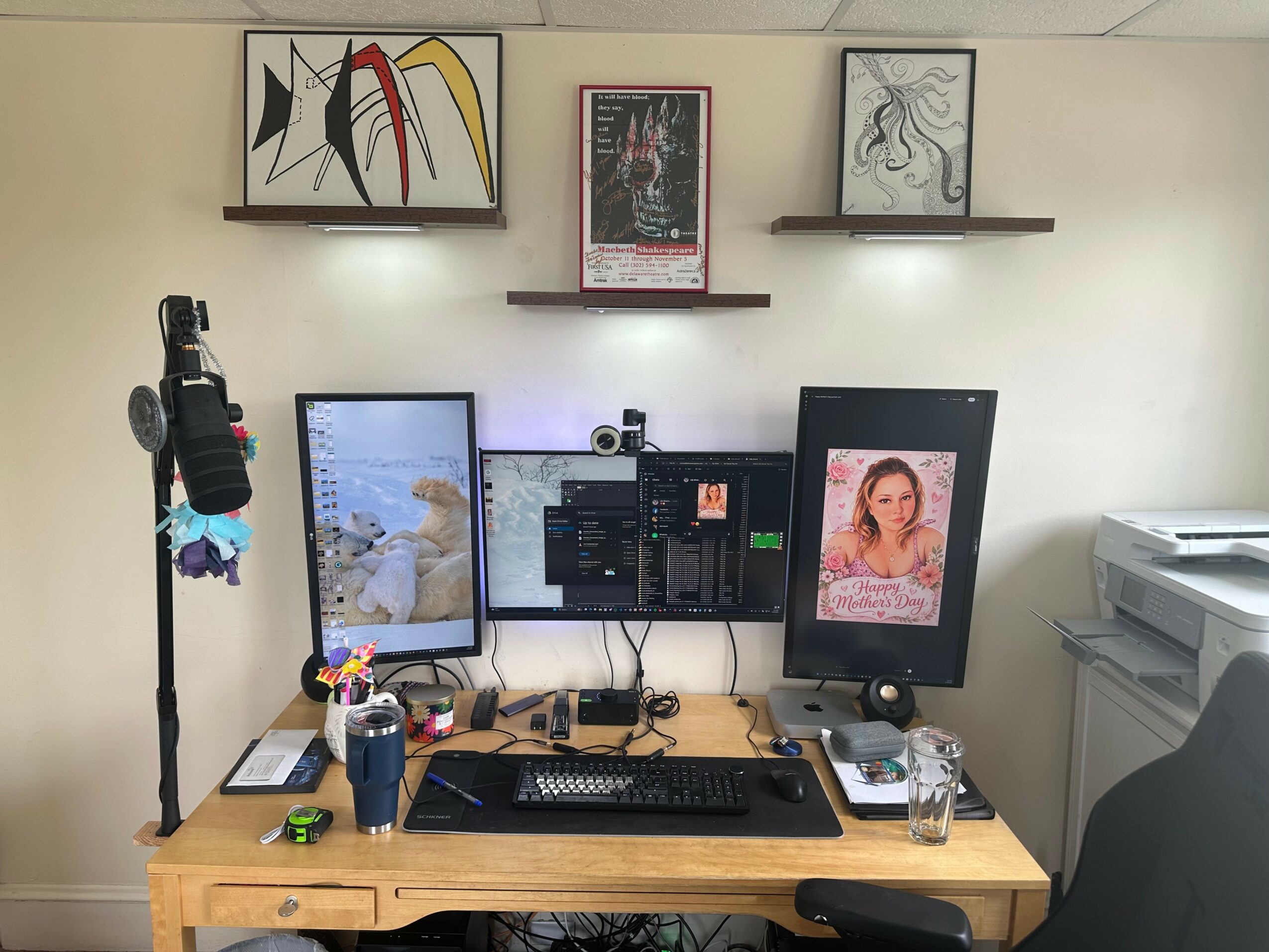 Office desk with two monitors, a keyboard, mouse, and various items, against a wall with three framed artworks and a poster.