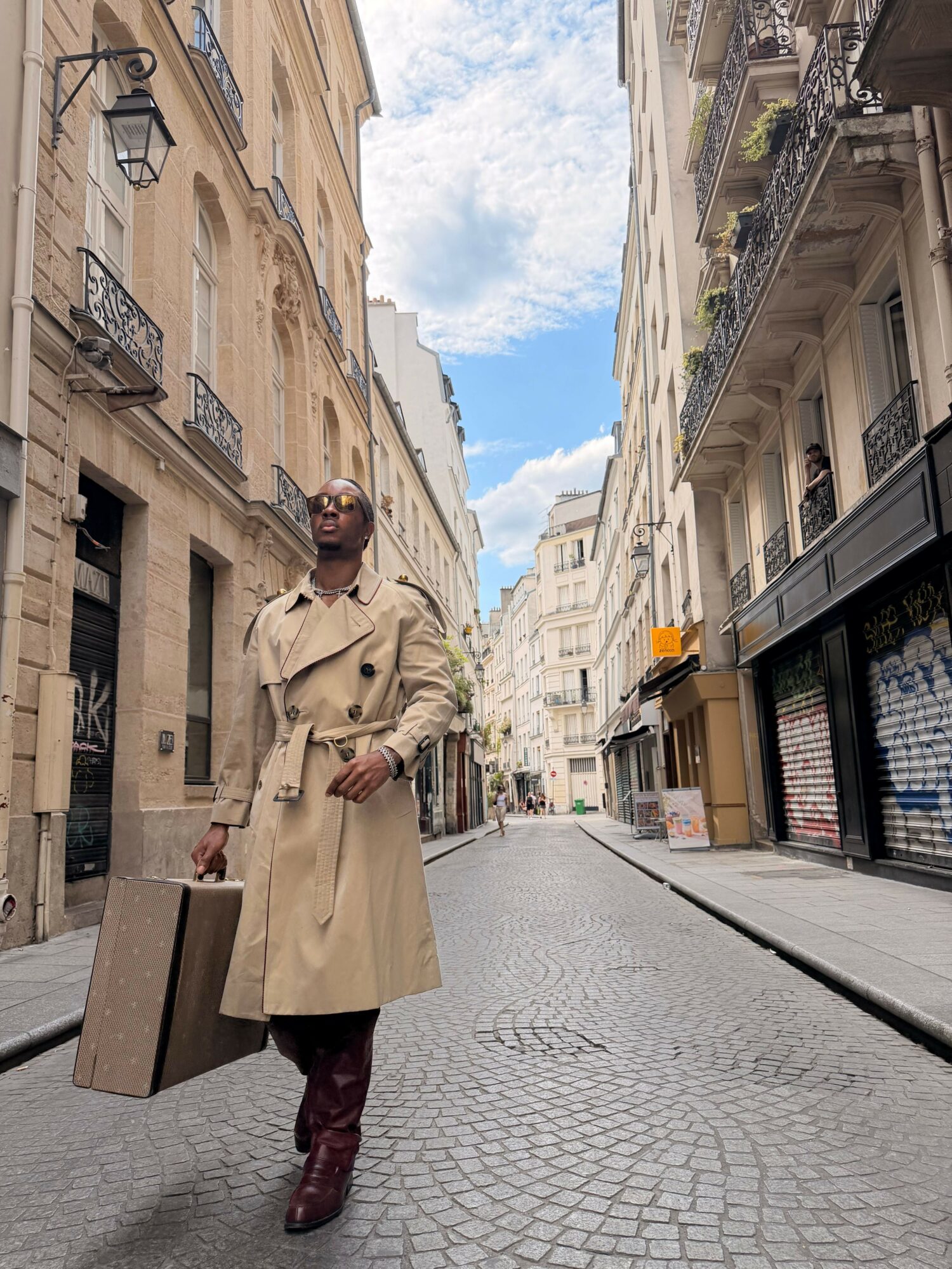 Person in beige coat and dark boots walking on a city street with buildings and a cloudy sky.