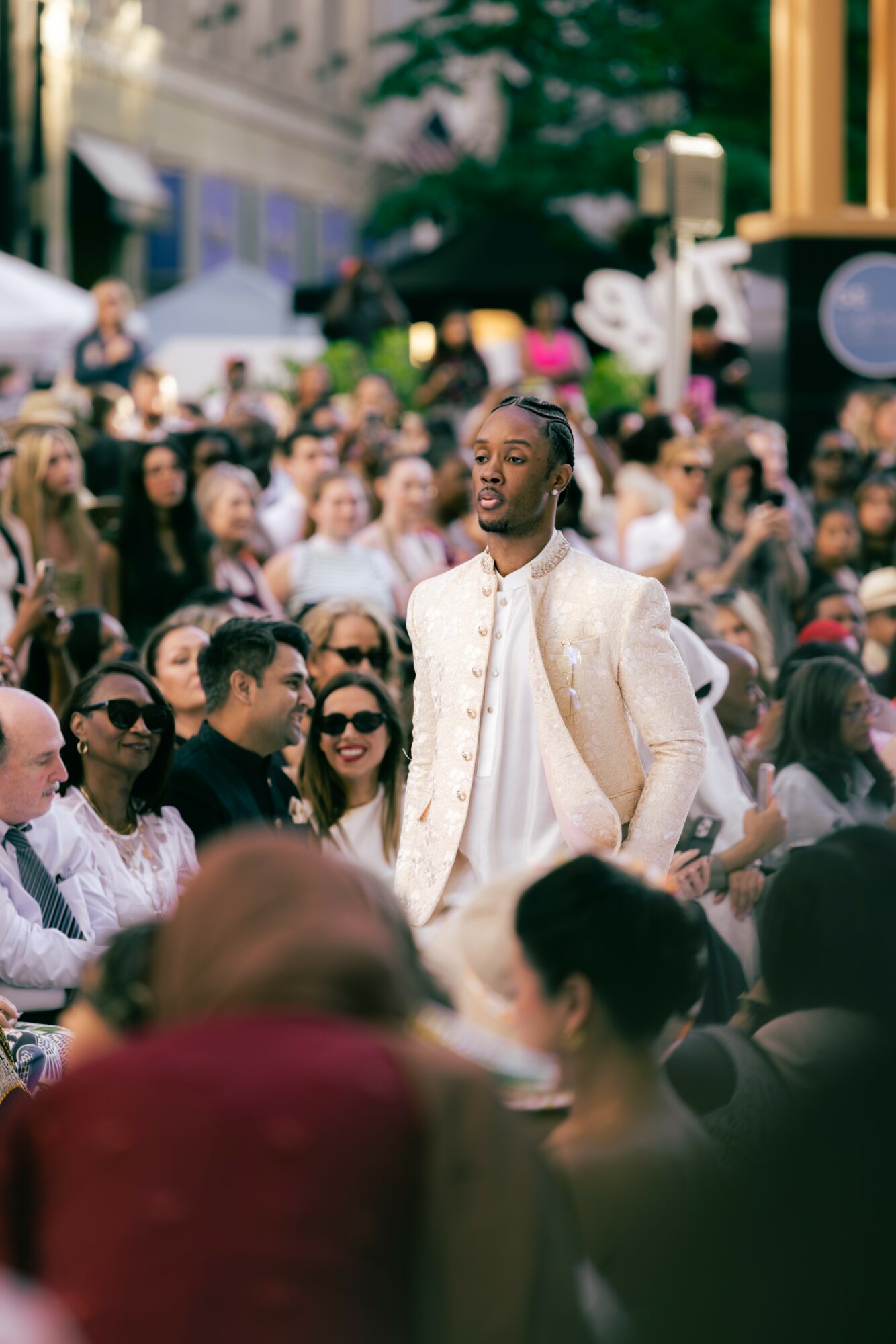 A man in traditional attire stands among a crowd at an outdoor event, with people smiling and wearing sunglasses.