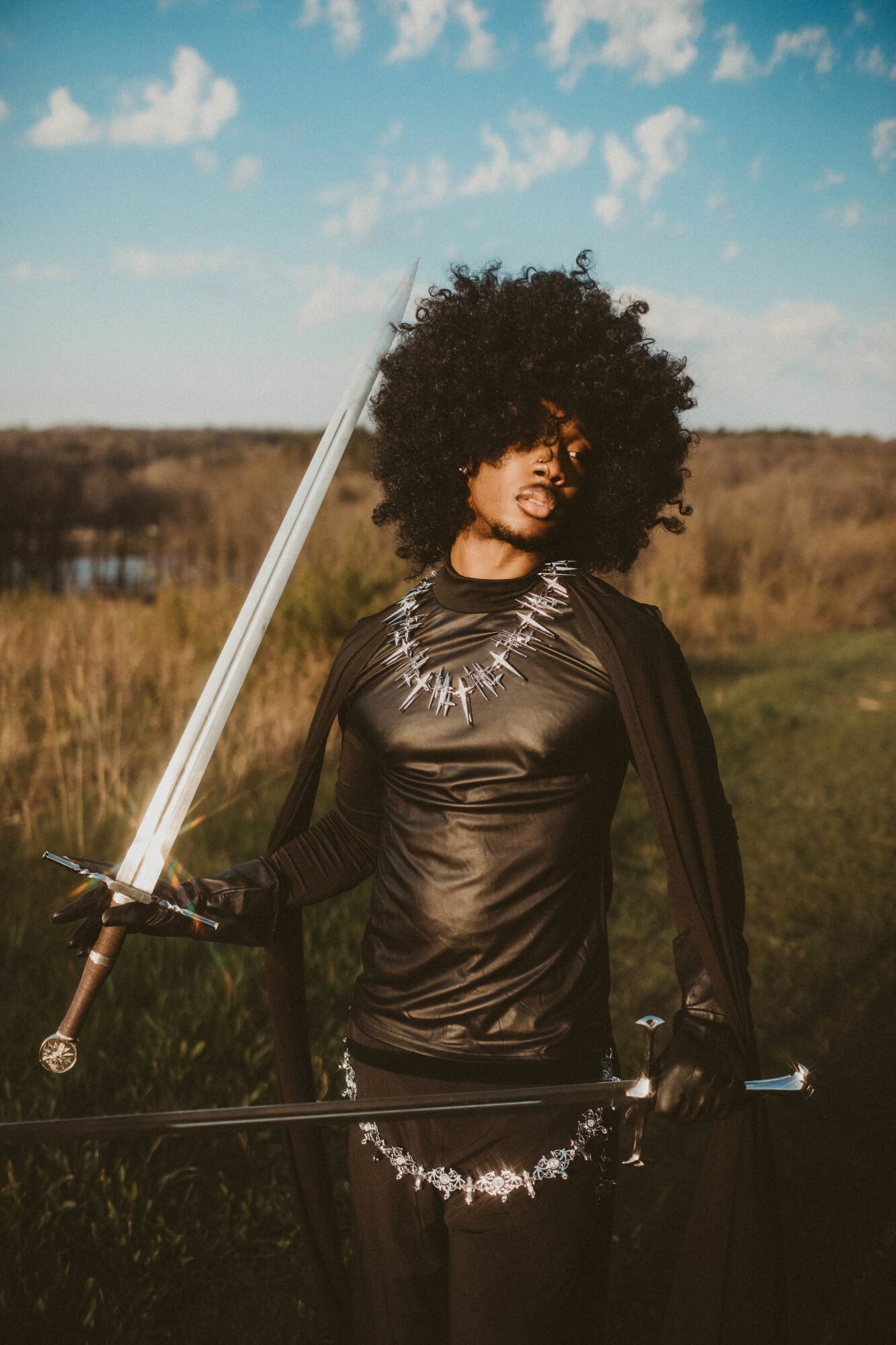 Person with curly hair holding a sword outdoors, wearing a black outfit with metallic details, in a grassy field under a blue sky.