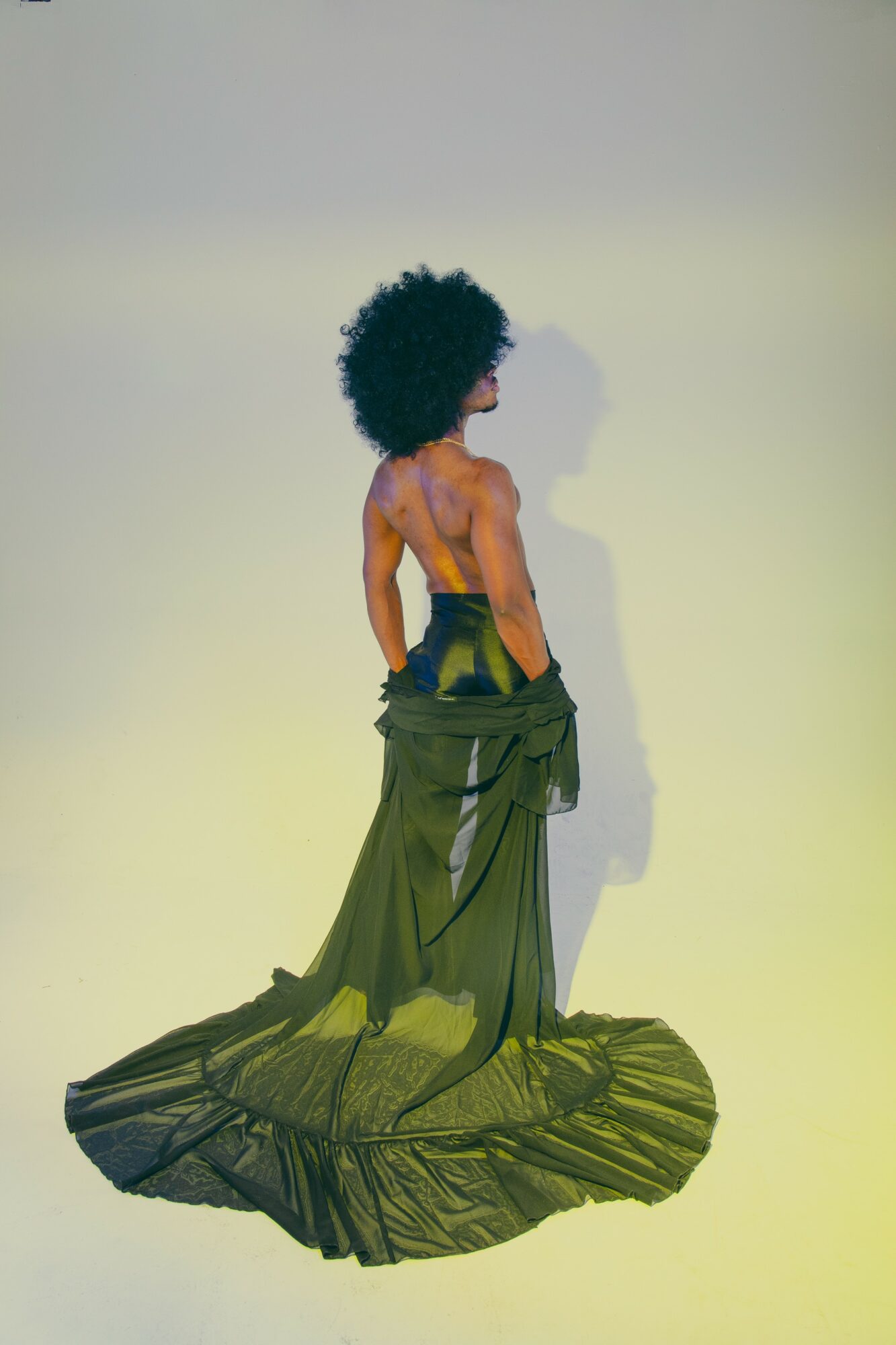 Person with curly hair wearing a long, flowing green dress, standing sideways against a plain background.