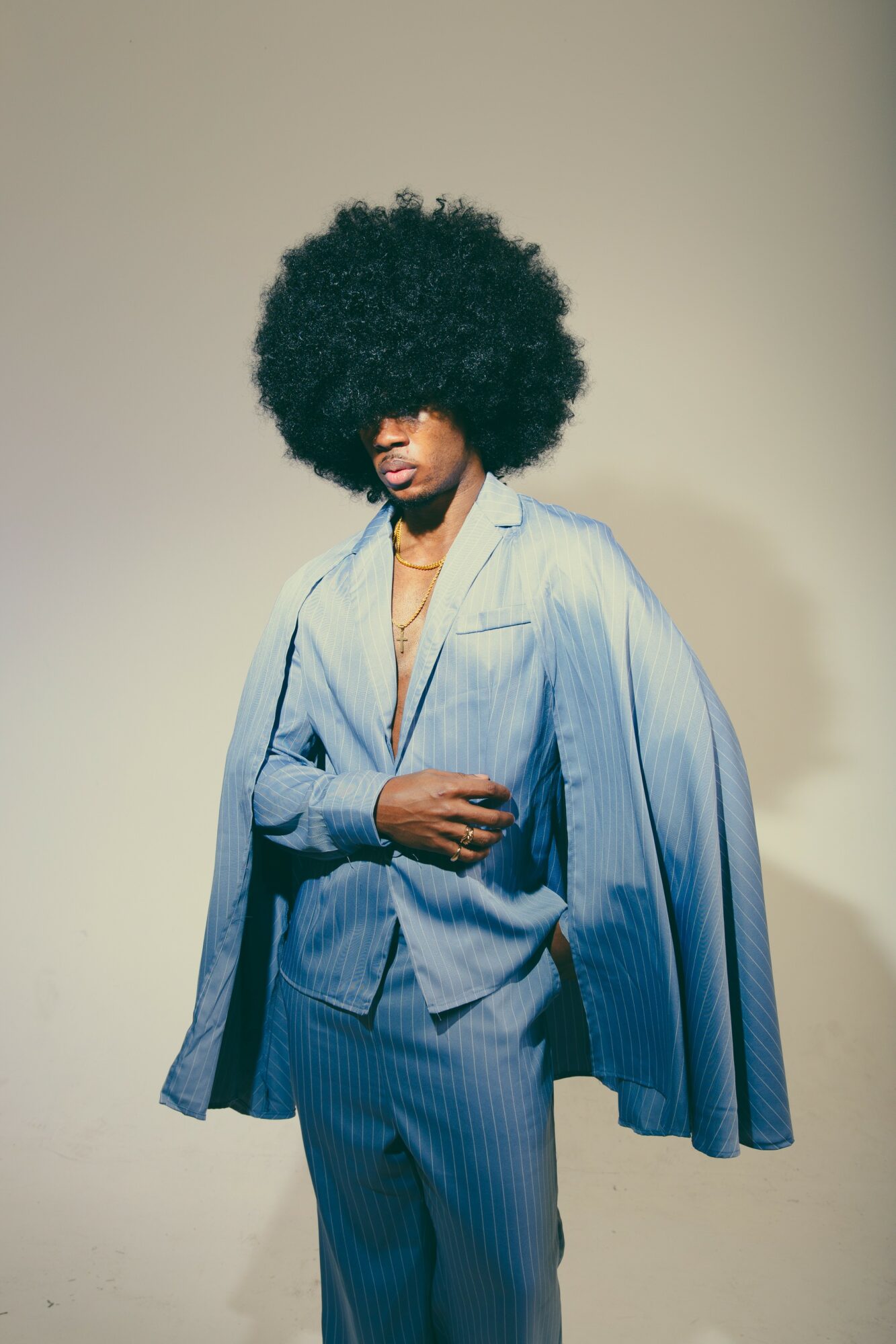 Person with large curly hair wearing a light blue suit and cape, standing against a plain background.
