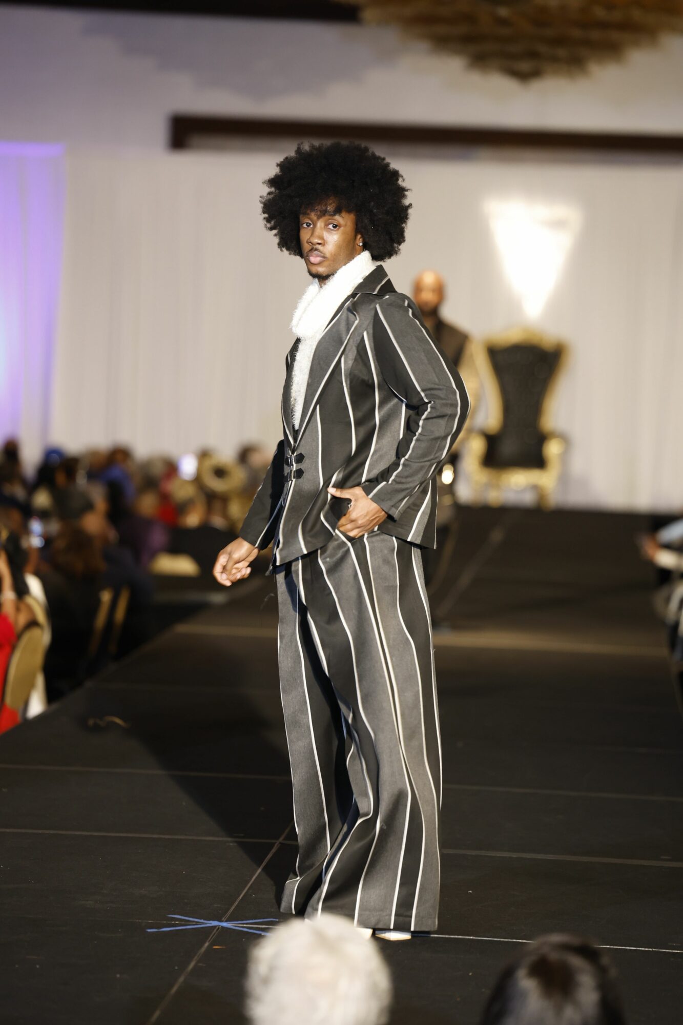 Person in a striped suit walking on a runway with an audience in the background.