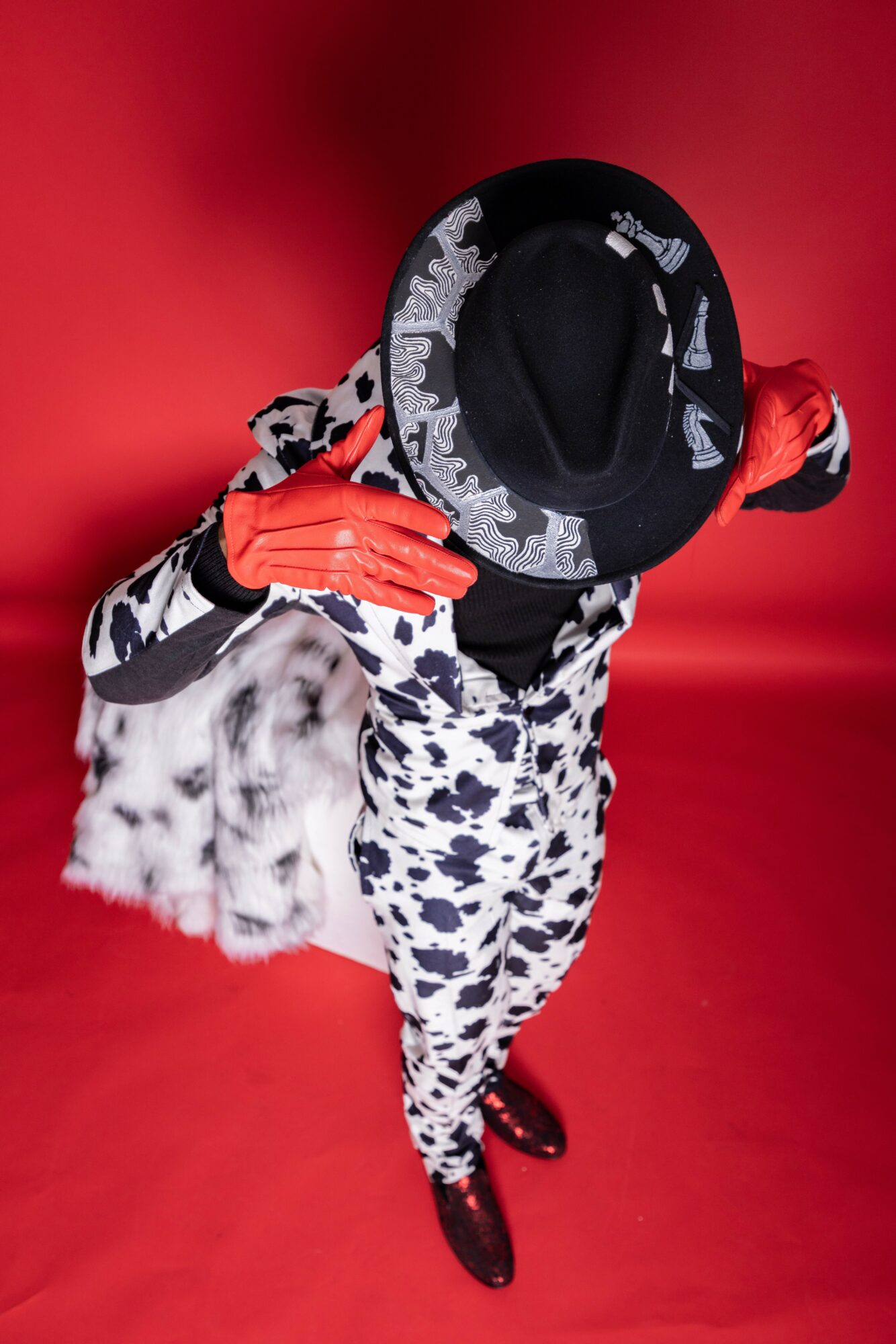 Person dressed in patterned clothing, holding a fur bag, wearing a hat and gloves, against a red background.