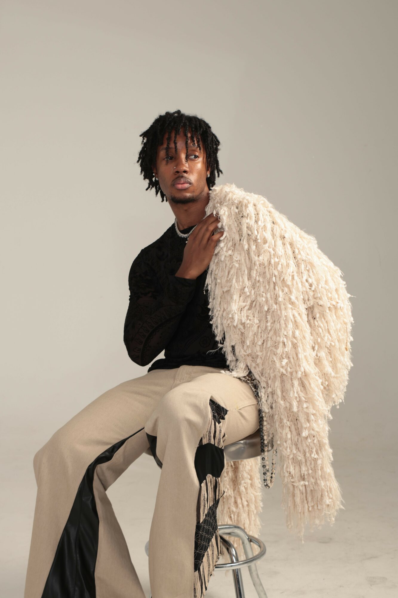 Person with dreadlocks lying on a fluffy white rug, wearing a black top and beige pants, against a plain background.