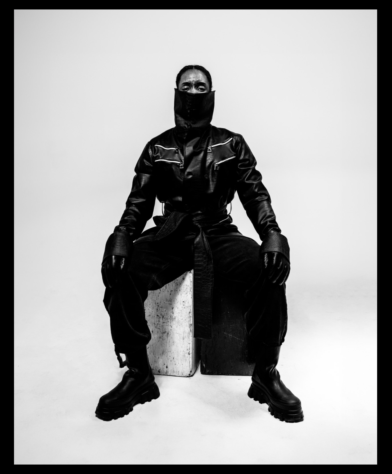 Person wearing a face mask, leather jacket, and gloves, sitting on a block in a studio with a plain background.