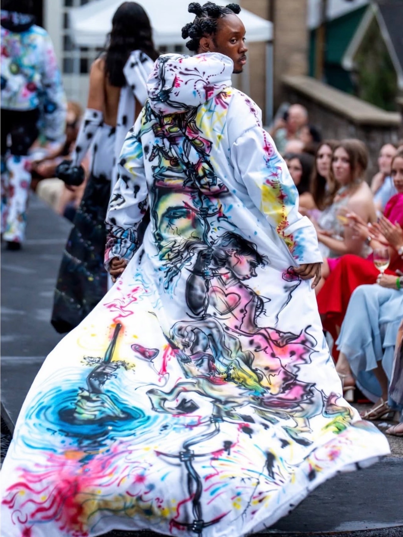 Model walking on runway wearing a colorful, abstract-patterned gown with a long flowing train, audience watching.