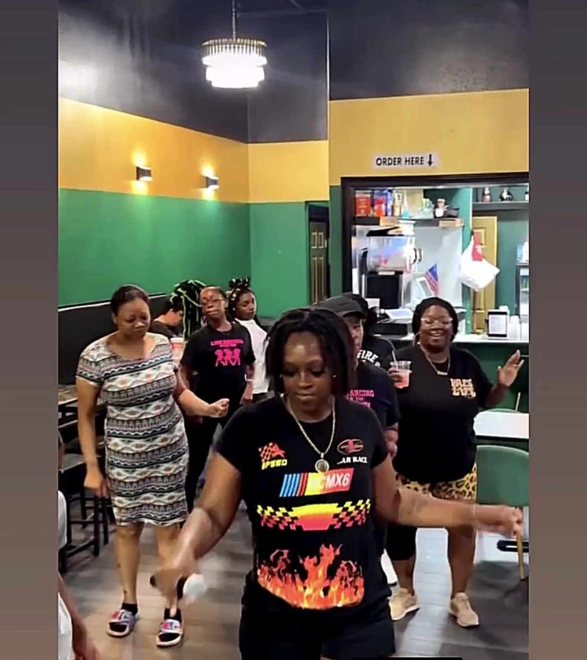 Group of women dancing indoors with colorful walls and a kitchen area in the background.