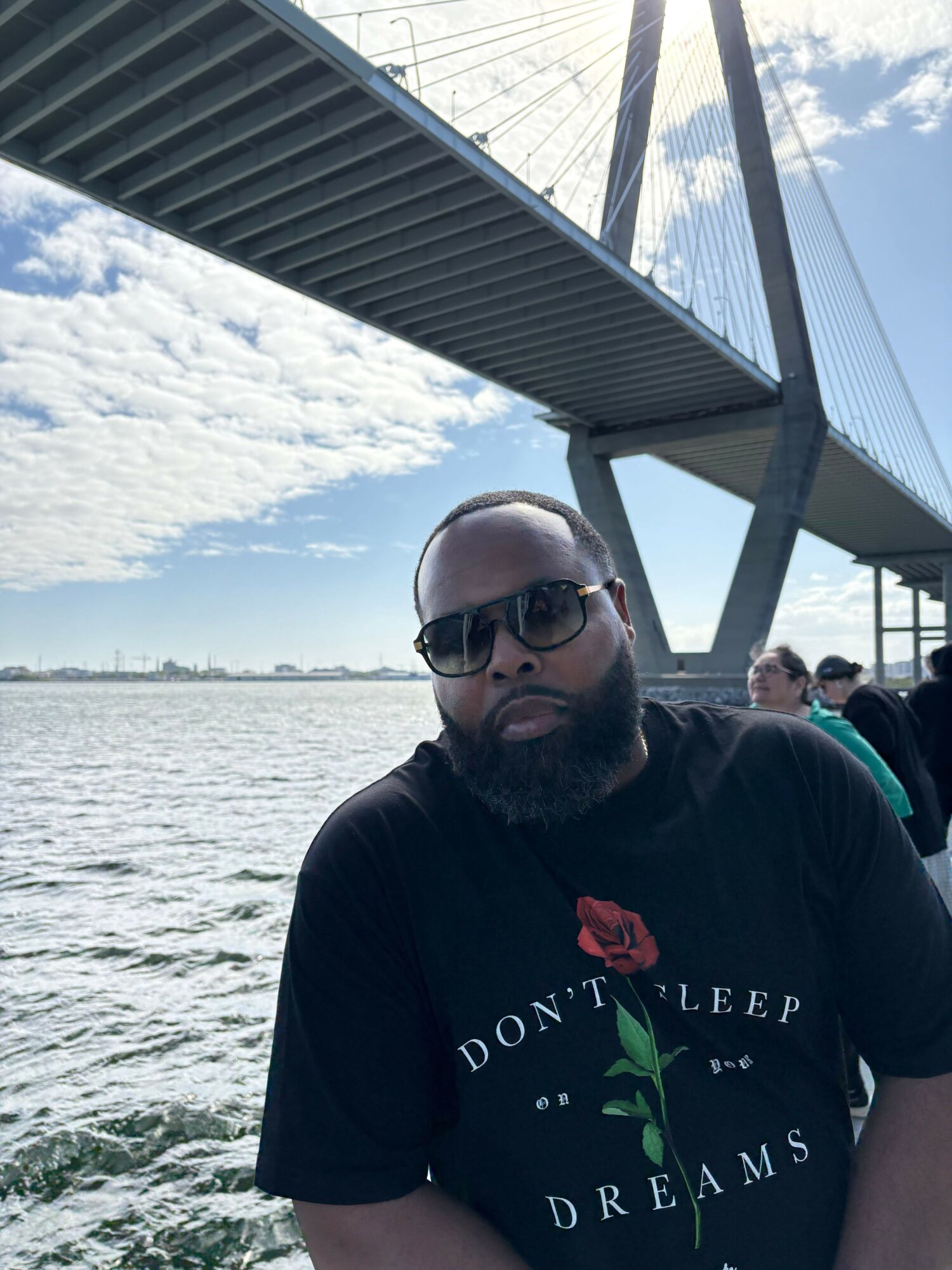 Person with sunglasses and bald head standing near water with a bridge overhead.