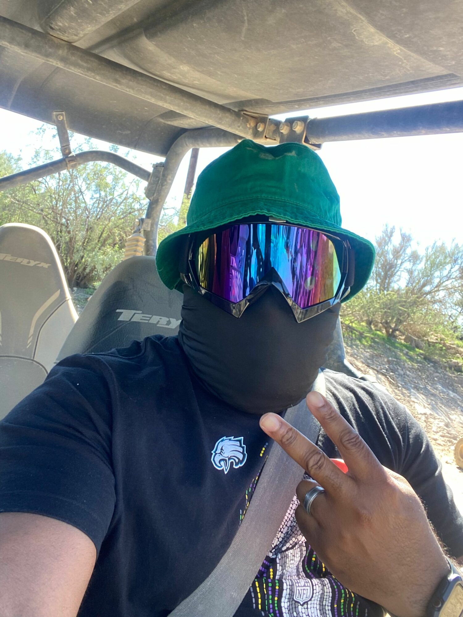 Person wearing a black shirt and gloves, holding colorful sunglasses, pointing with their right hand, outdoors with trees and sky.