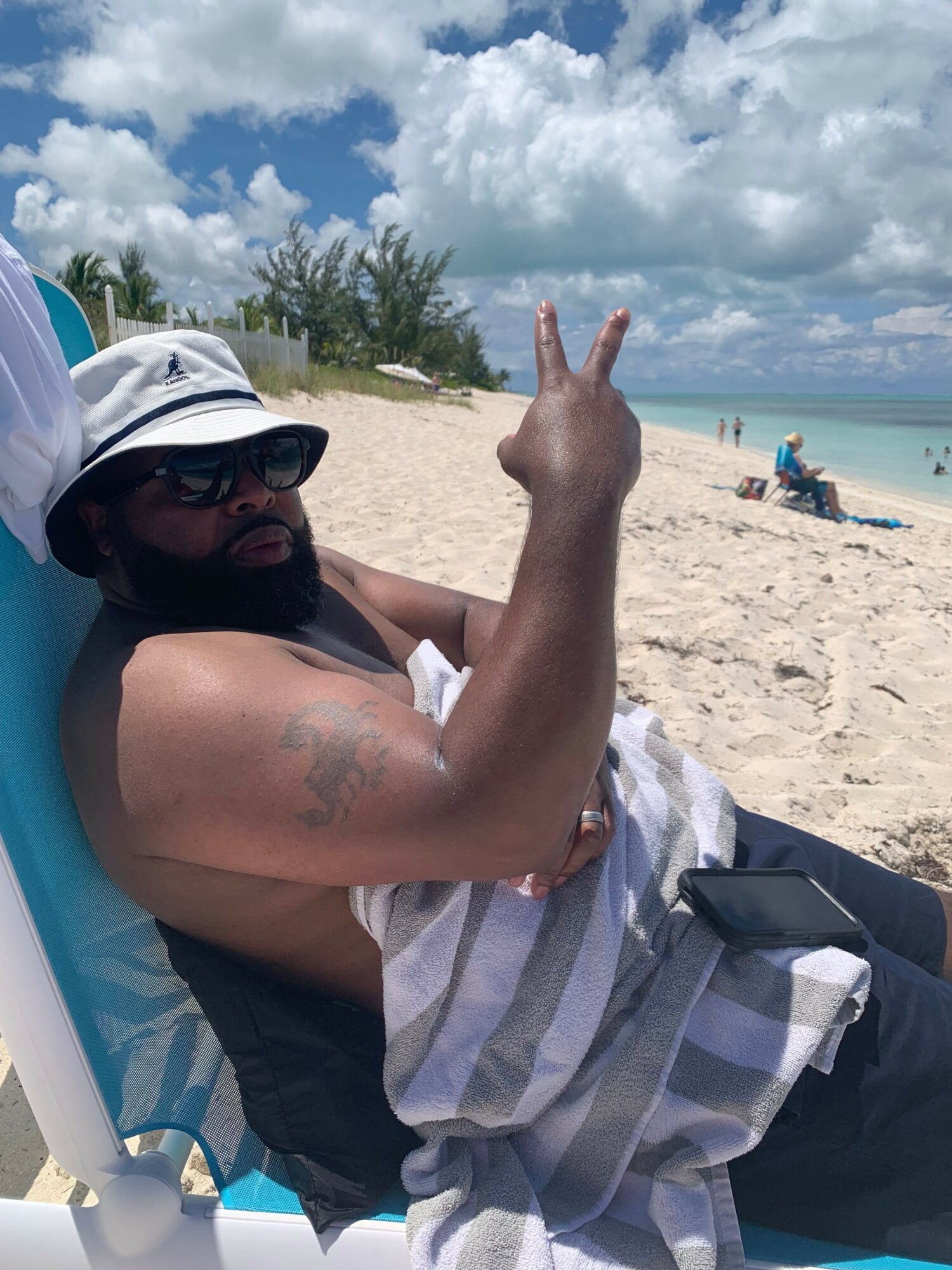 Person lying on a beach chair making a peace sign, wearing sunglasses and a hat, with sand and sky in background.