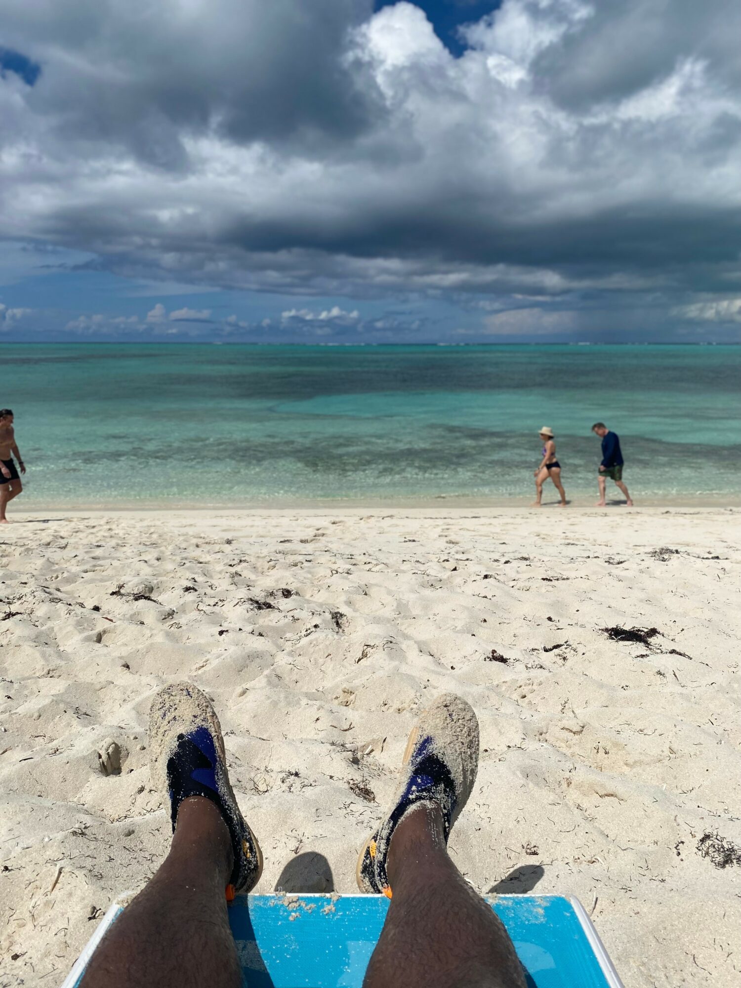 Person sitting on a beach chair with legs stretched out, wearing sneakers, facing the ocean with cloudy sky.