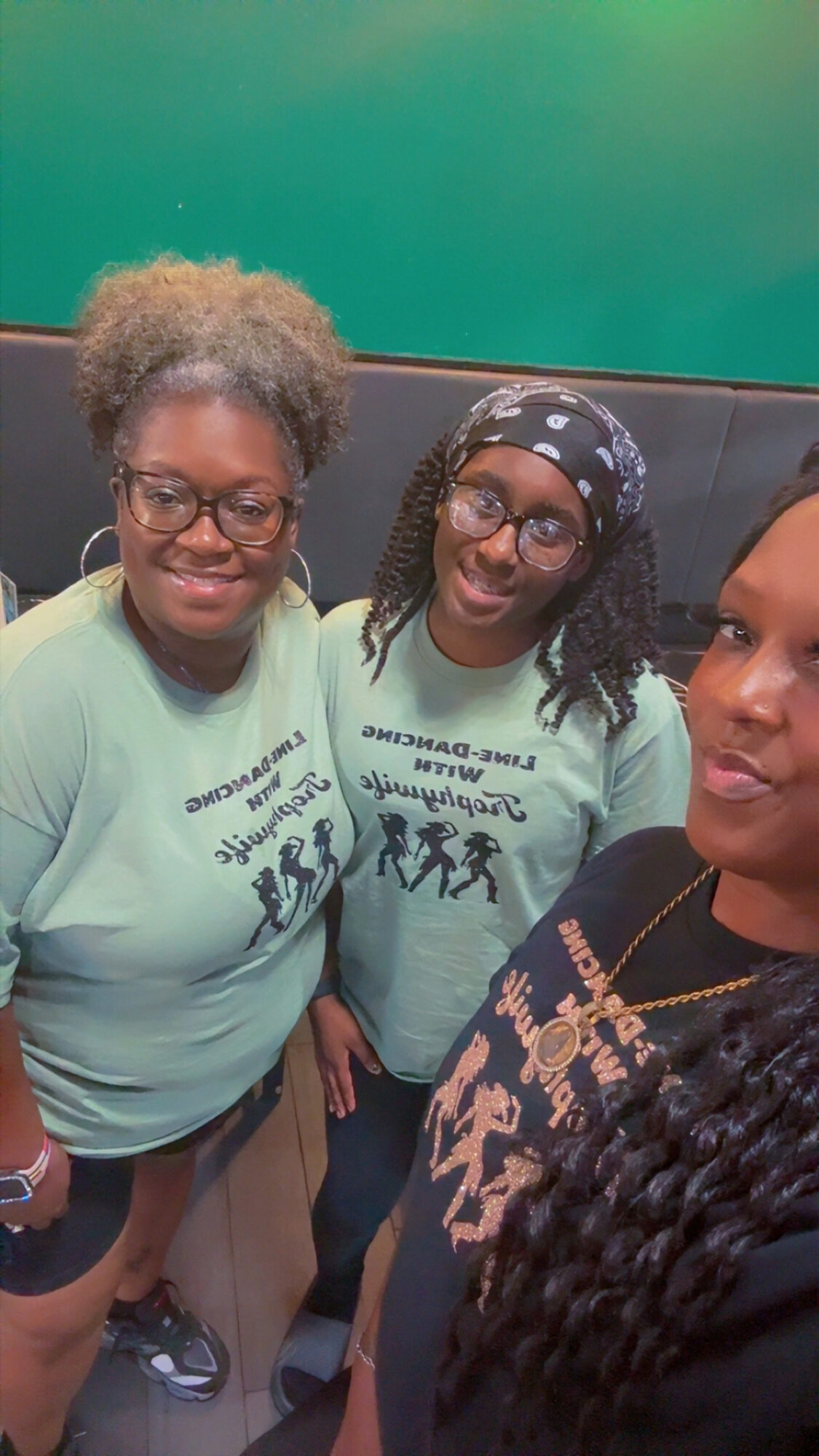 Three people smiling, two women wearing glasses and light green shirts, one woman with long braided hair and a patterned top.