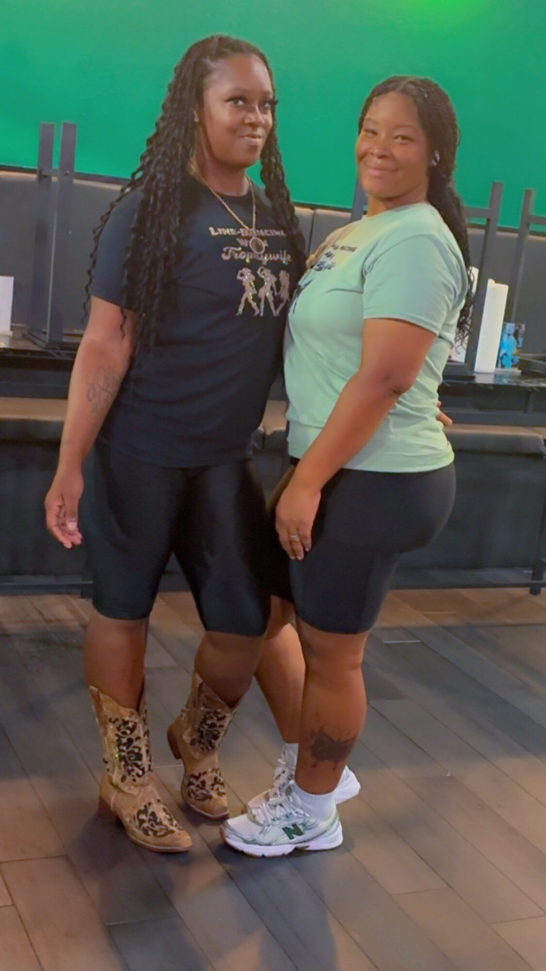Two women standing side by side indoors, smiling, with a green background and black counter behind them.