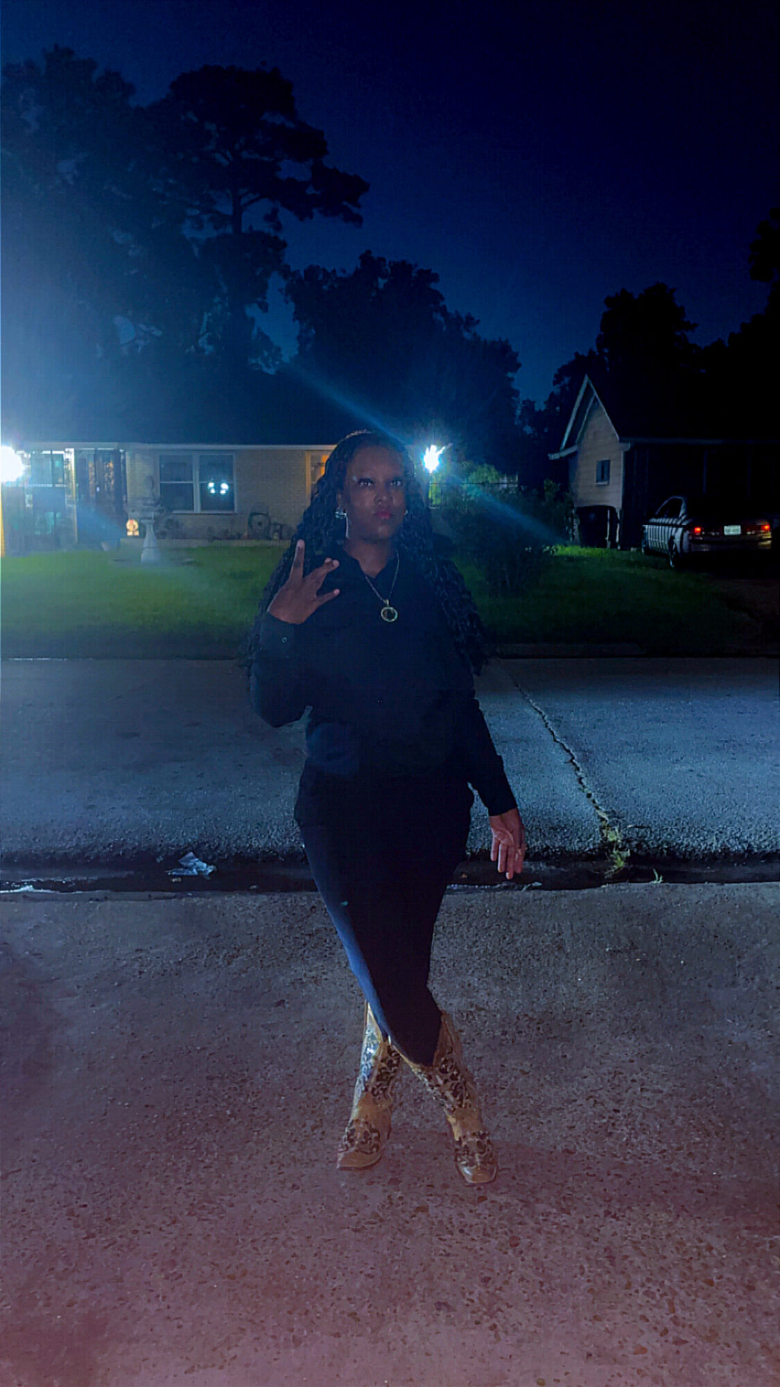 Person standing on street at night, making a peace sign with their right hand, with houses and trees in background.