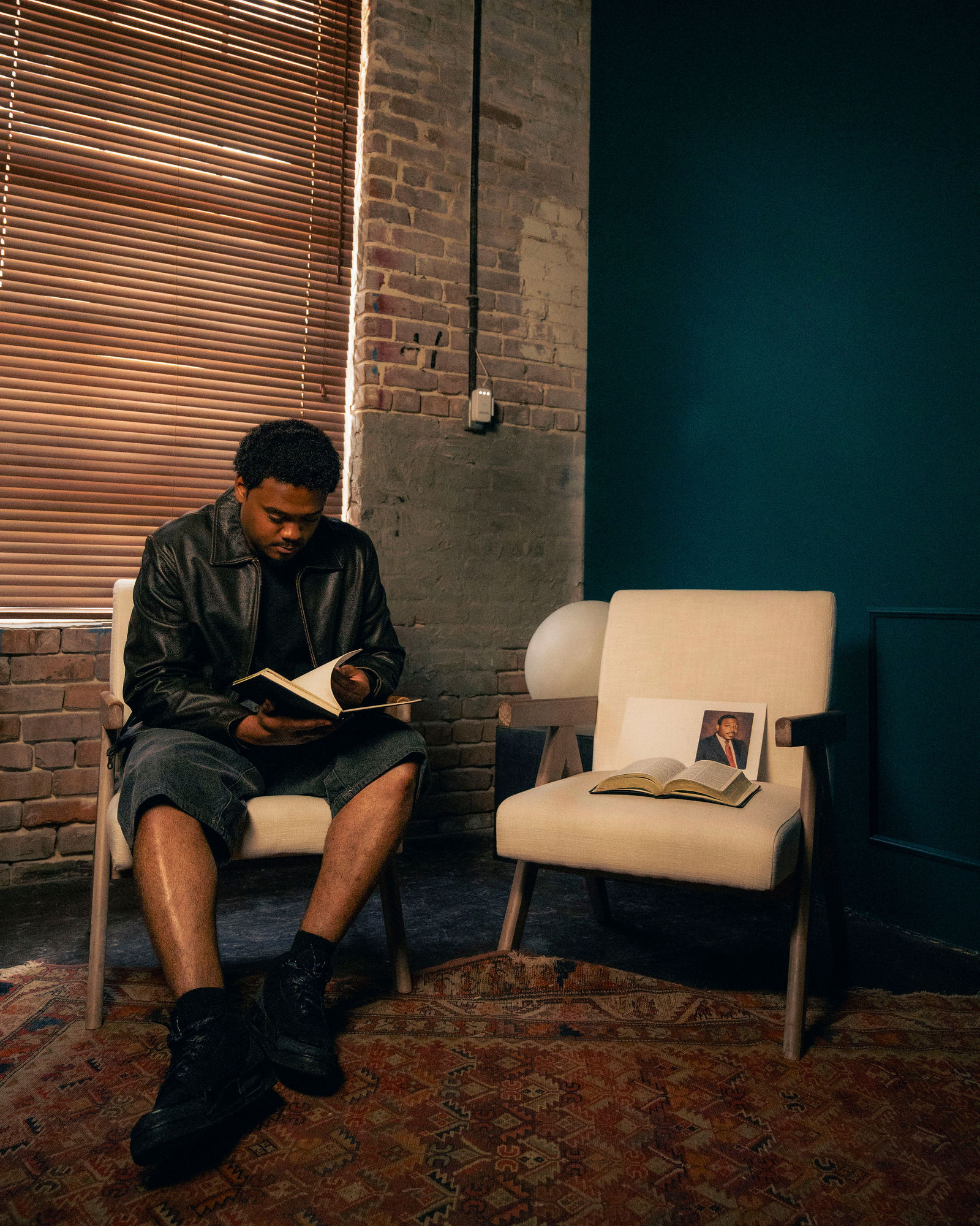 Man sitting on a chair reading a book in a room with brick wall, wooden blinds, and modern furniture.
