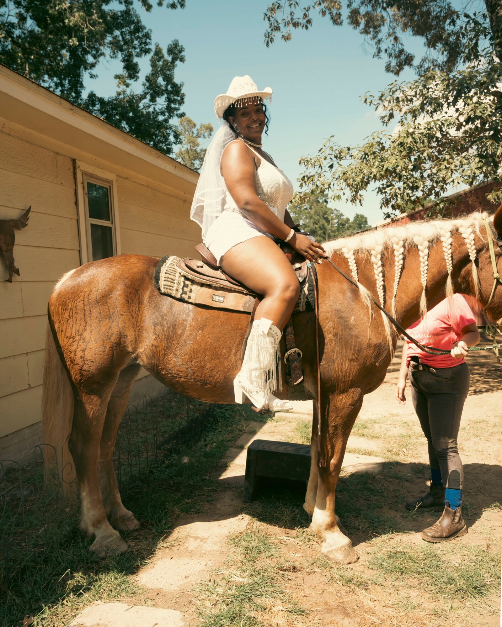 Woman wearing a white hat and outfit, sitting on a brown horse outdoors, with trees and a building in the background.