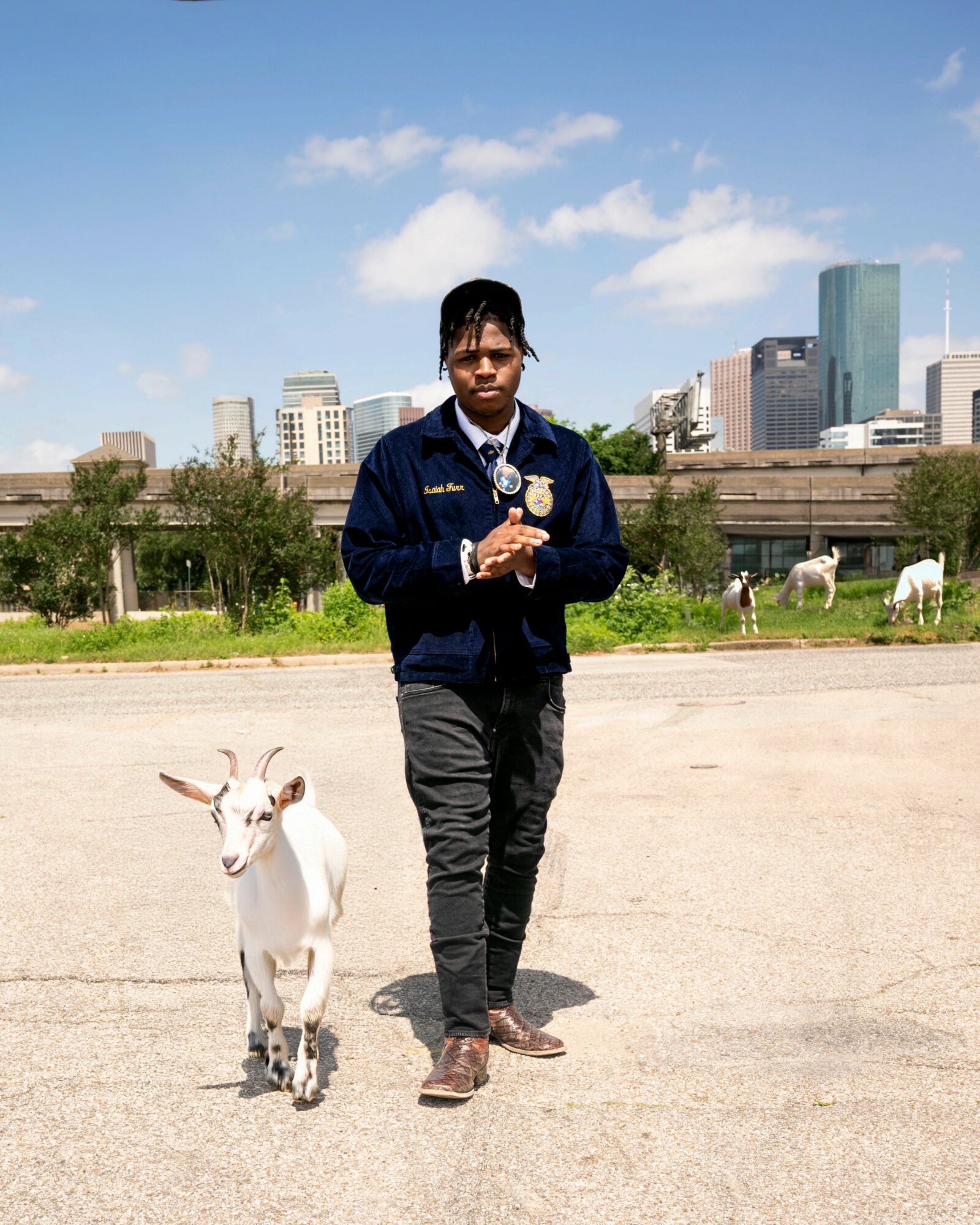 Young man walking outdoors with a white goat, city skyline in background, blue sky, wearing dark jacket and pants.