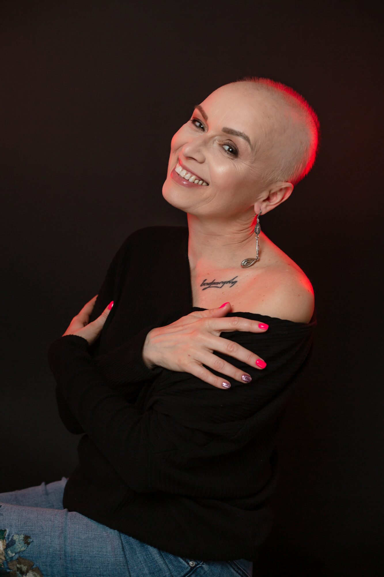 Smiling person with a shaved head, wearing a black top, crossed arms, and a tattoo on the shoulder, against a dark background.