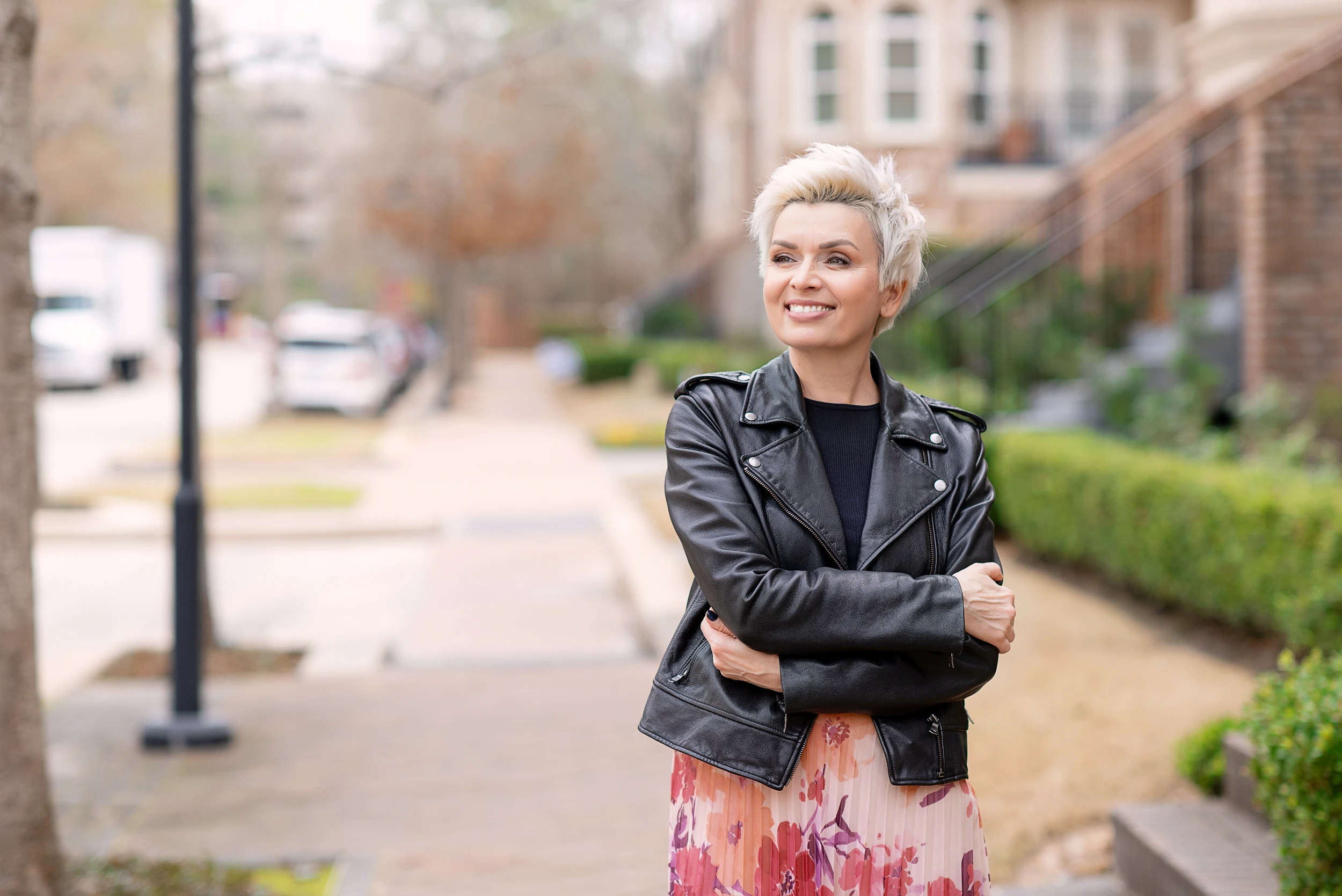 Smiling woman with short blonde hair wearing a black leather jacket and patterned skirt, standing outdoors on a sidewalk.
