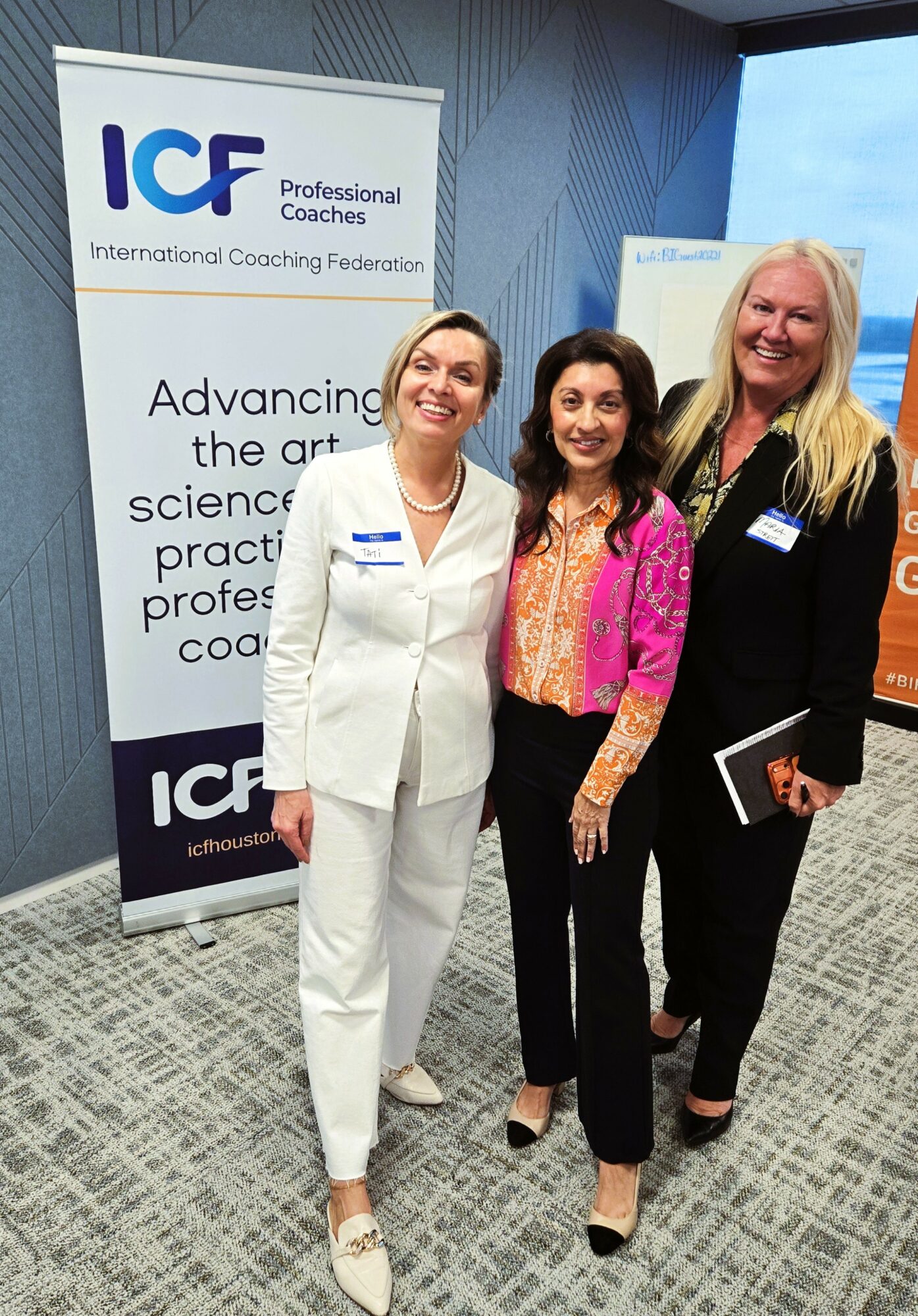 Three women standing together indoors, smiling, with a banner and window in the background.