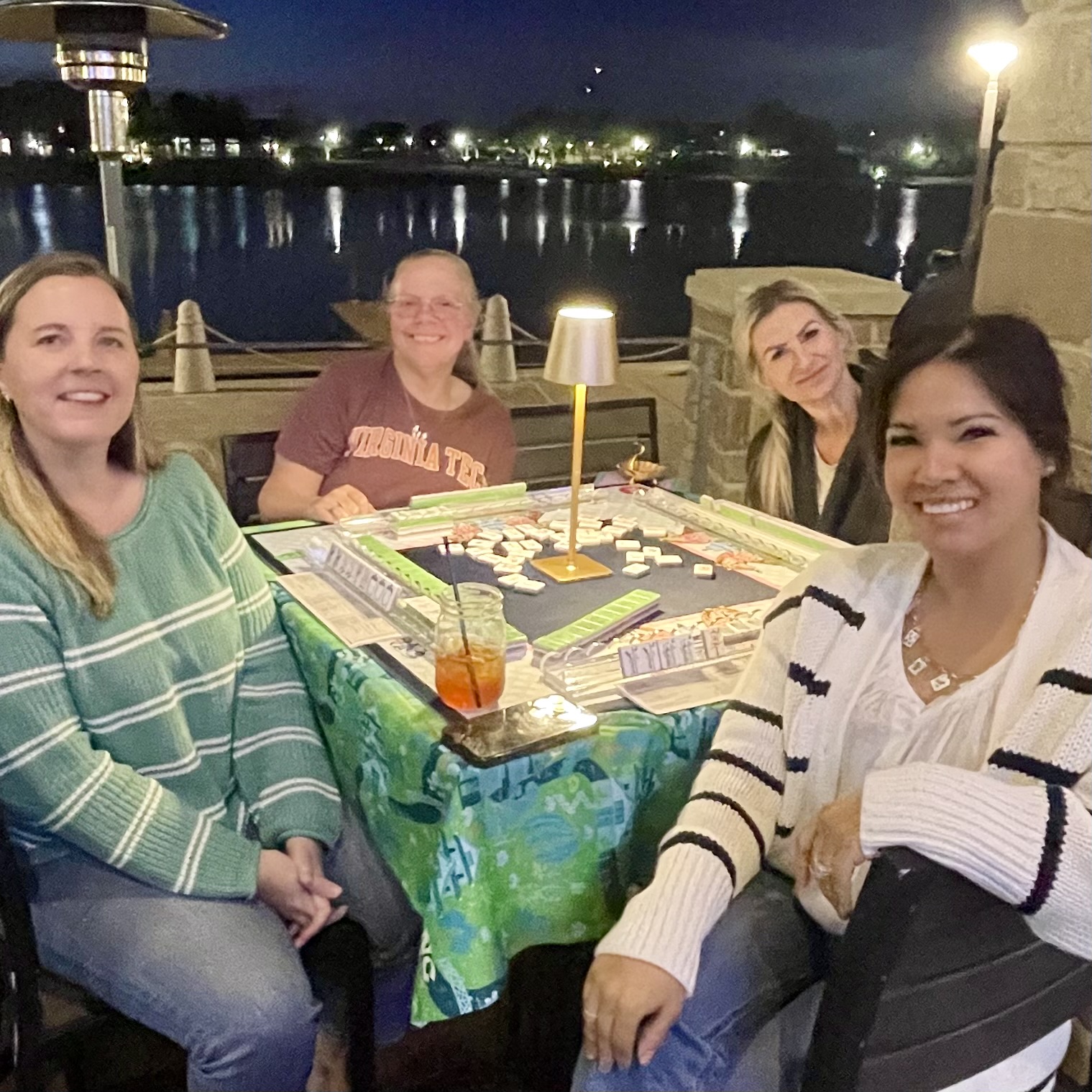 Four women sitting around a table outdoors at night, with a body of water and city lights in the background.