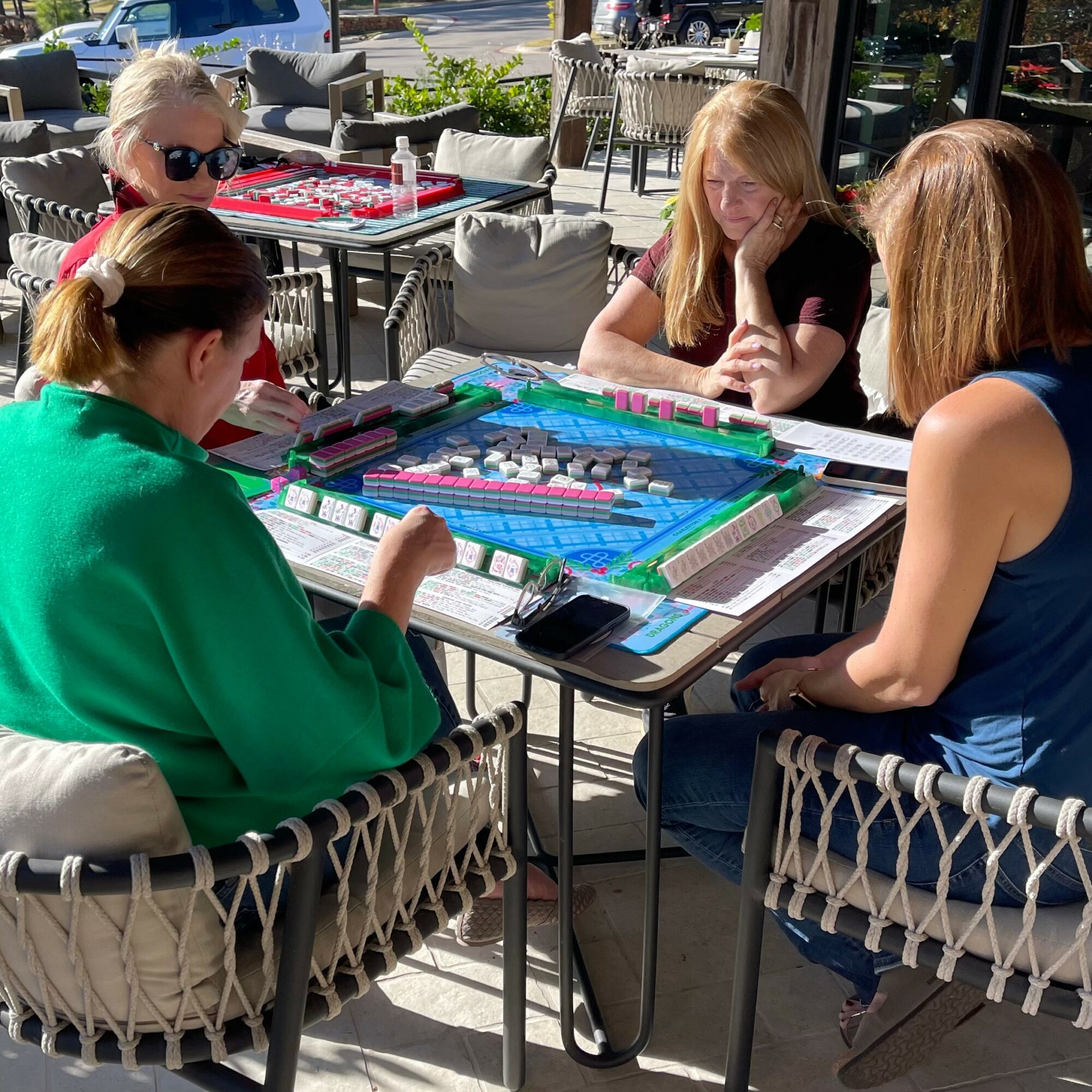 Four women playing a board game outdoors at a table with sunlight and chairs around, some wearing sunglasses.