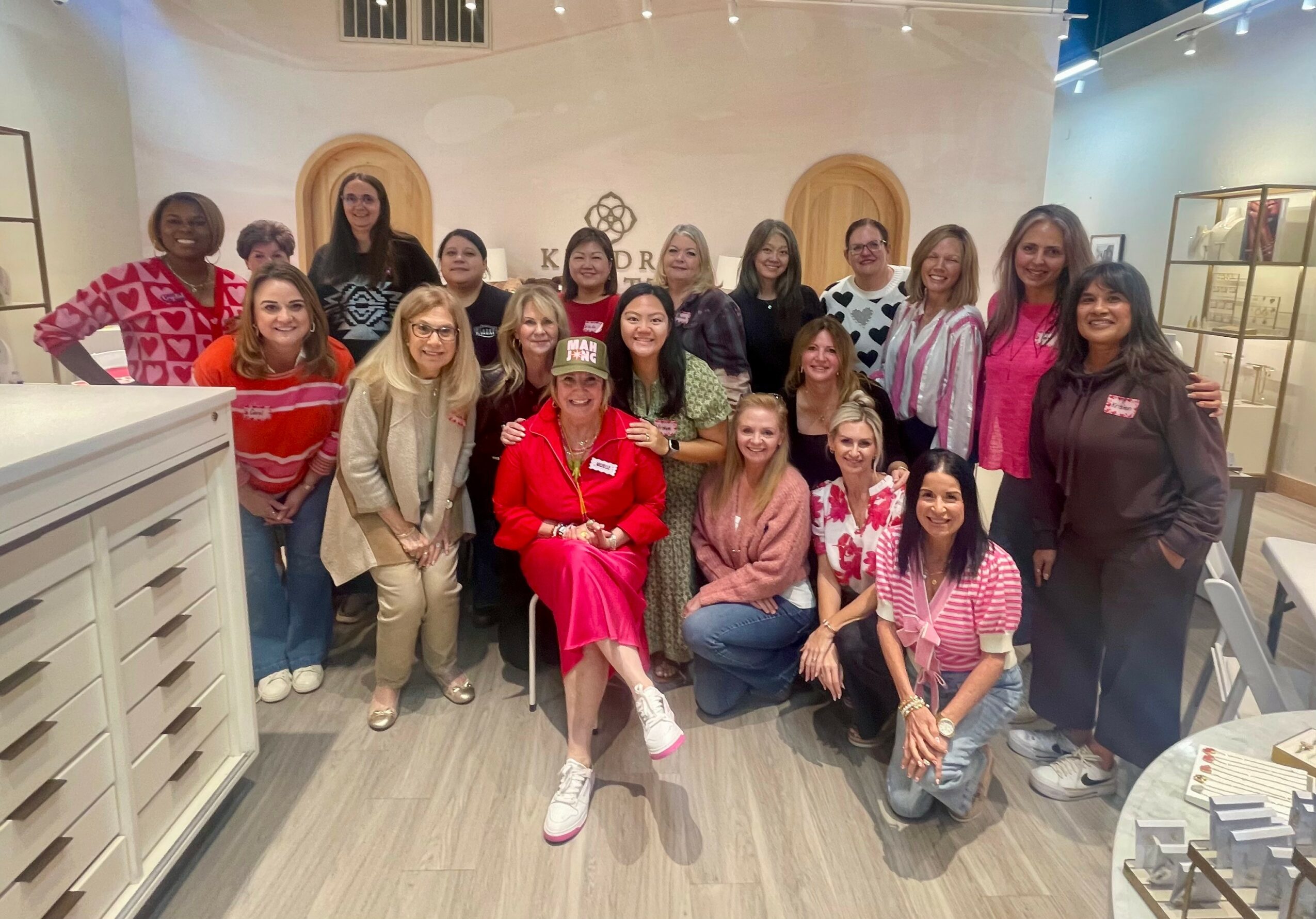 Group of 16 women and girls posing indoors, some sitting and some standing, in a well-lit room with wooden decor.
