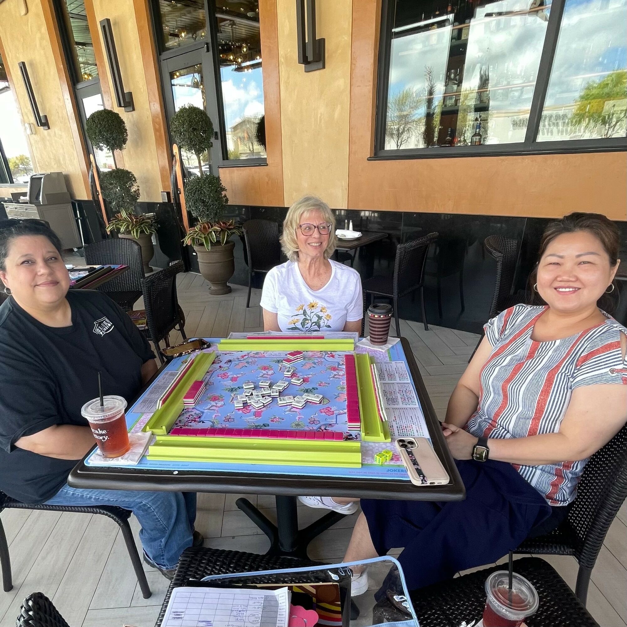 Three women sit around a table playing a game with colorful pieces, in a bright indoor setting with large windows.