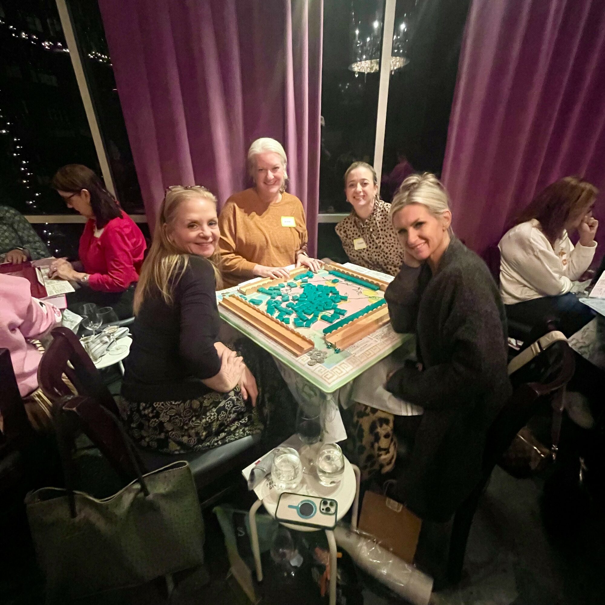 Four women sit around a table with a large decorated cake, smiling at the camera, in a room with purple curtains.