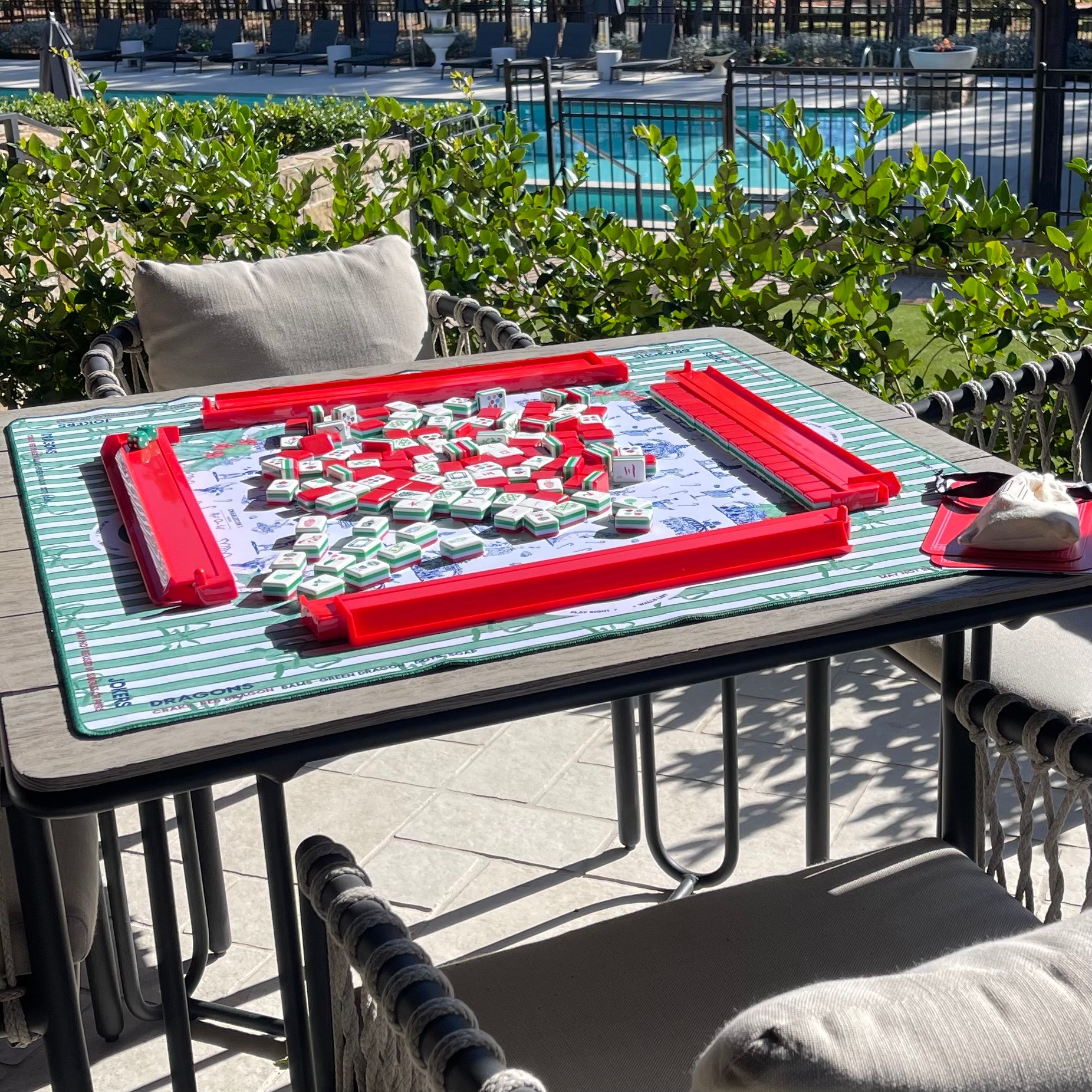 Table with a glass top, surrounded by chairs with cushions, outdoors near greenery and a pool, with red and white game pieces on the table.