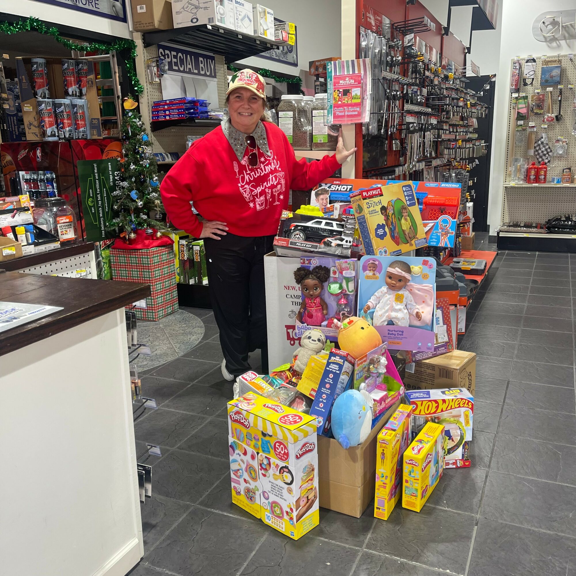 Woman in Christmas sweater and hat standing next to a display of toys in a store, holiday decorations visible.