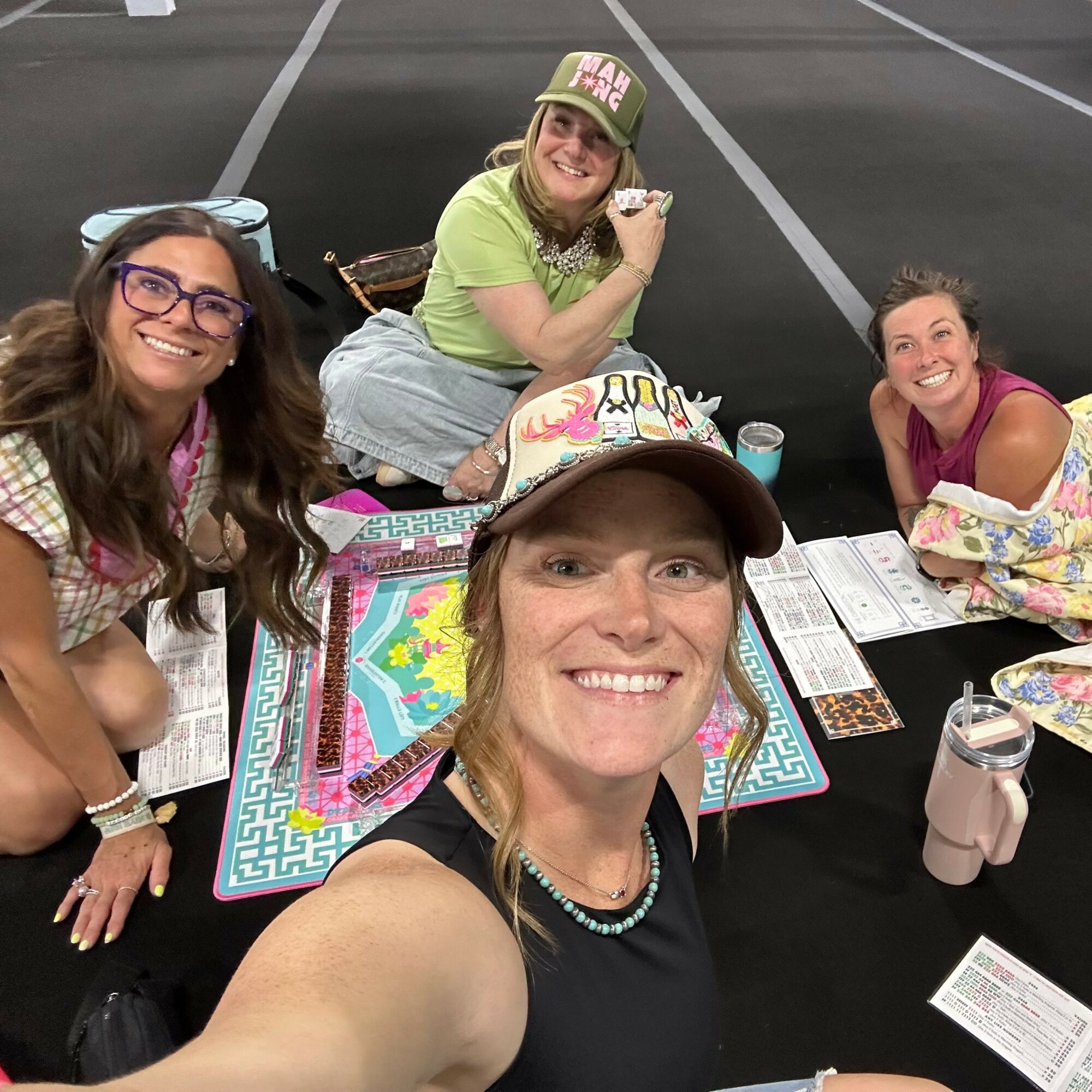 Four women sitting around a table playing a board game, smiling, with drinks and papers on the table.