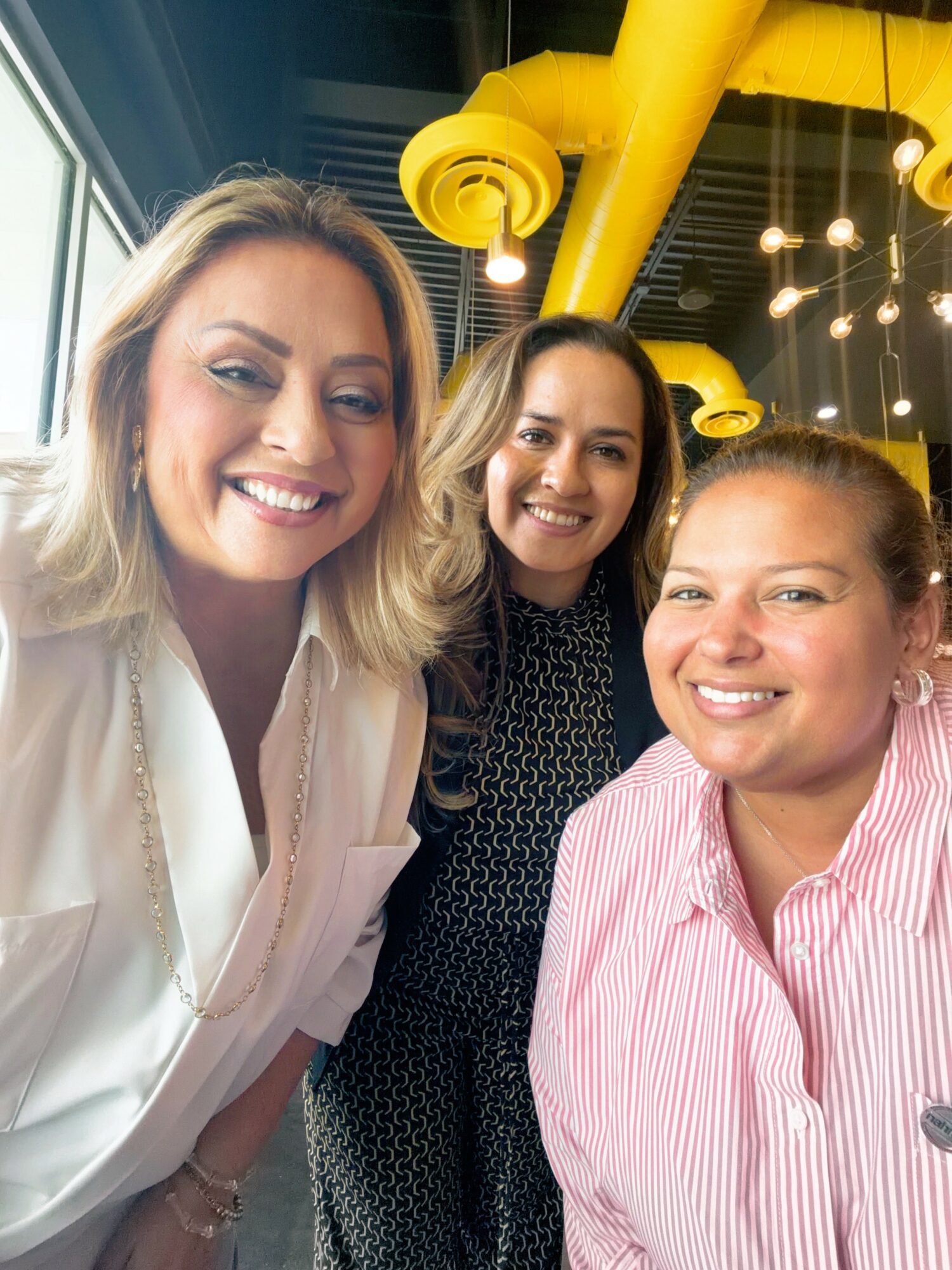 Three women smiling indoors with yellow ceiling pipes and hanging lights in the background.