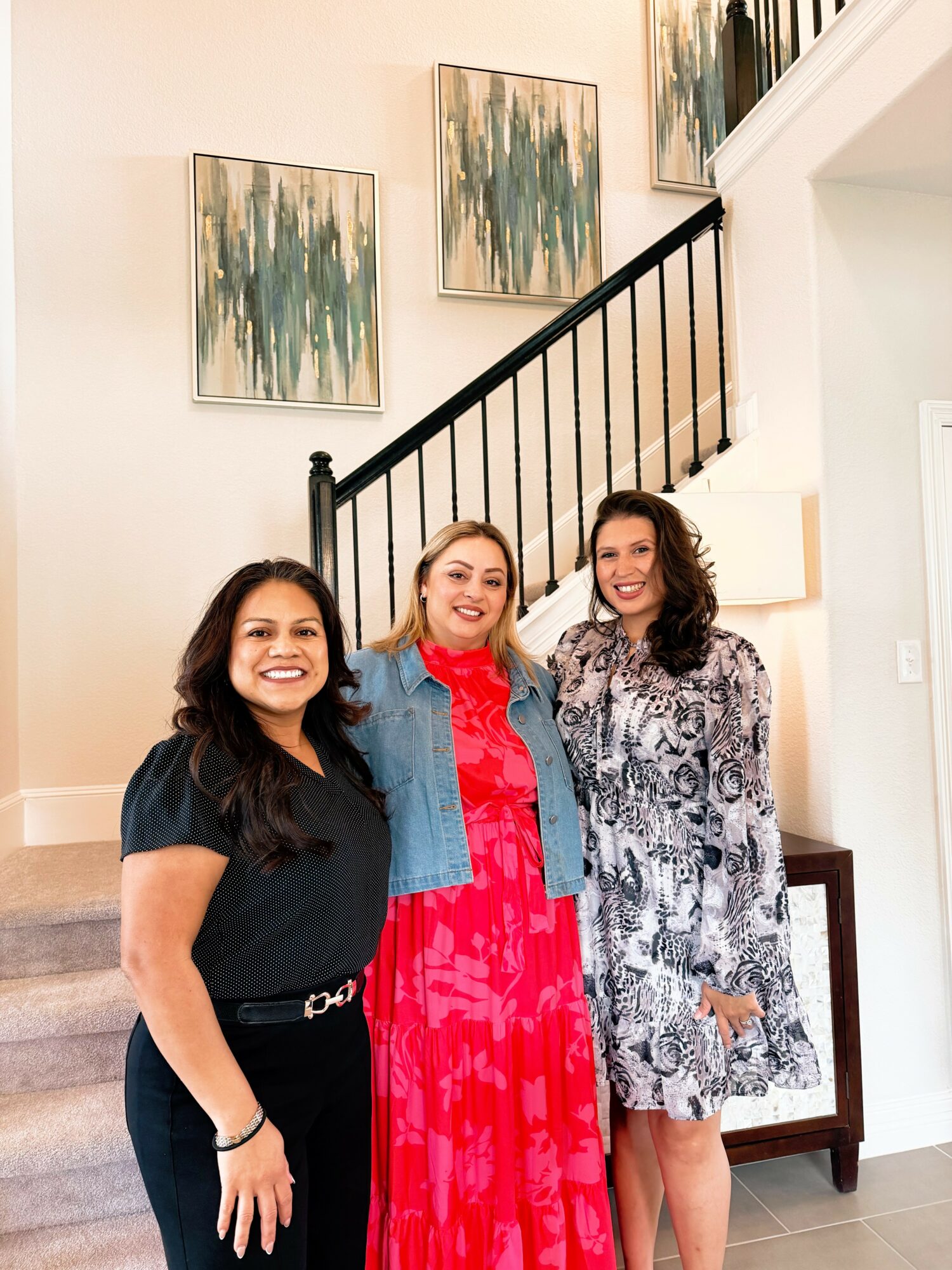 Three women standing indoors near a staircase, smiling, with abstract paintings on the wall behind them.
